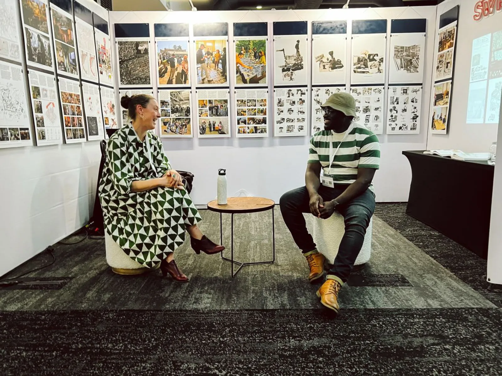 Zwei Personen sitzen in einer Ausstellung vor einer Wand mit zahlreichen Bildern und Texten. Sie unterhalten sich entspannt auf Hockern. Die Ausstellung thematisiert urbanes Design und soziale Realitäten in Afrika.