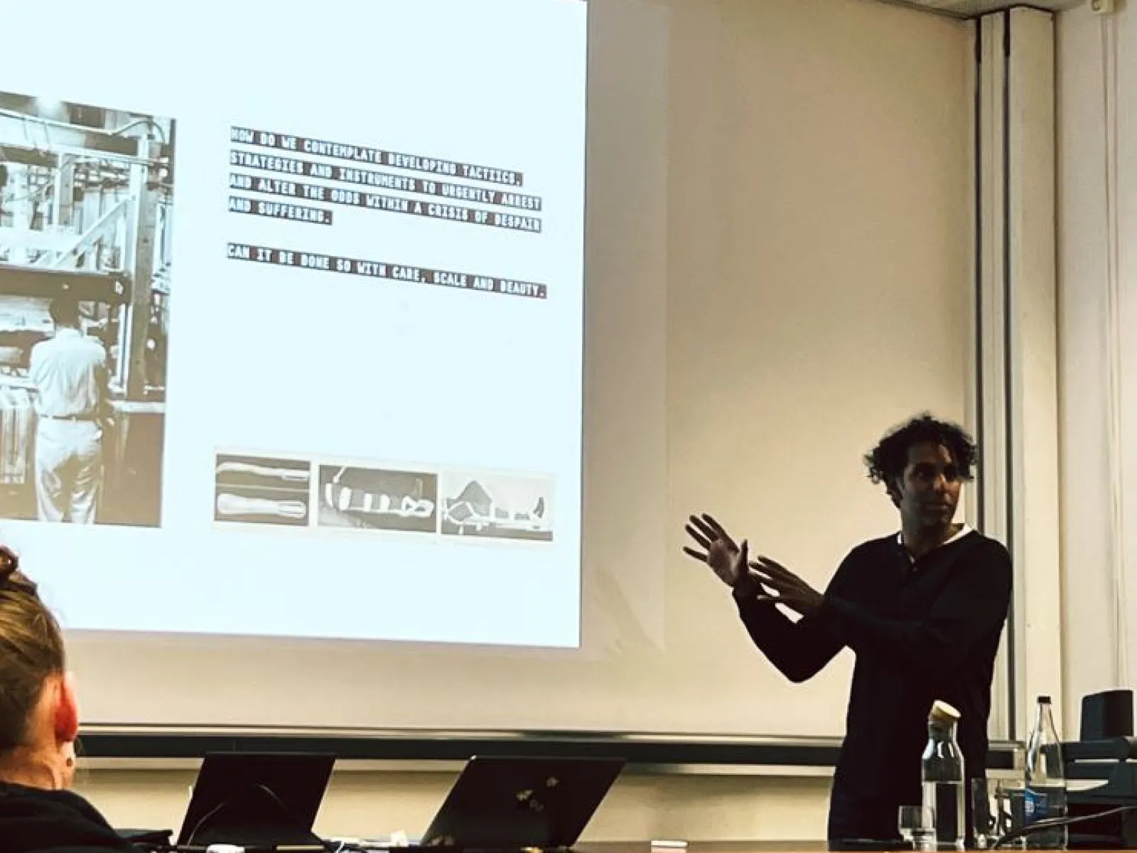 Ein Vortragender steht vor einer Projektionsleinwand und präsentiert Inhalte zu urbanem Design. Auf der Leinwand sind Text und Bilder zu sehen. Im Vordergrund sitzen Zuhörer. Der Raum wirkt modern und technisch ausgestattet.