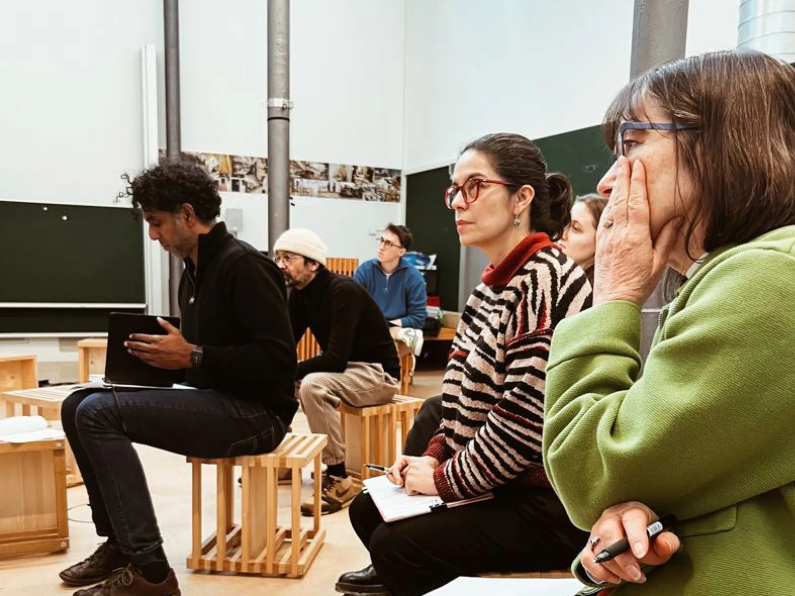 Eine Gruppe von Personen sitzt aufmerksam in einem Seminarraum. Sie sind auf Holzhockern platziert und scheinen einer Präsentation oder Diskussion zu folgen. Im Hintergrund sind Tafeln und Poster zu sehen. Die Szene vermittelt eine konzentrierte Lernatmosphäre.