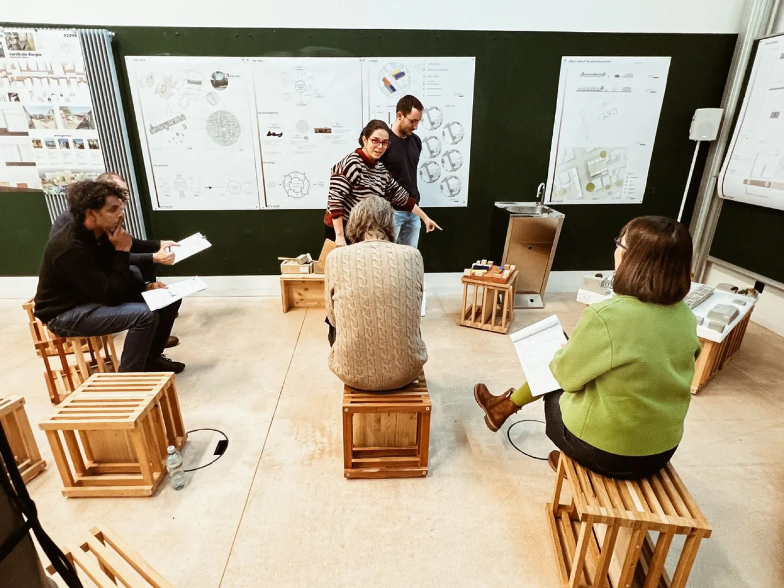 Eine Gruppe von sechs Personen sitzt auf Holzkisten in einem Raum mit Plakaten an der Wand. Zwei Personen stehen und diskutieren, während die anderen Notizen machen. Der Raum wirkt wie ein Workshop oder eine Präsentation.