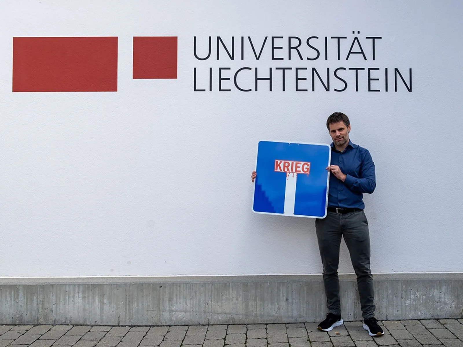 Ein Mann steht vor einer Wand mit dem Logo der Universität Liechtenstein und hält ein Verkehrsschild mit der Aufschrift „Krieg“ in einer Sackgasse.
