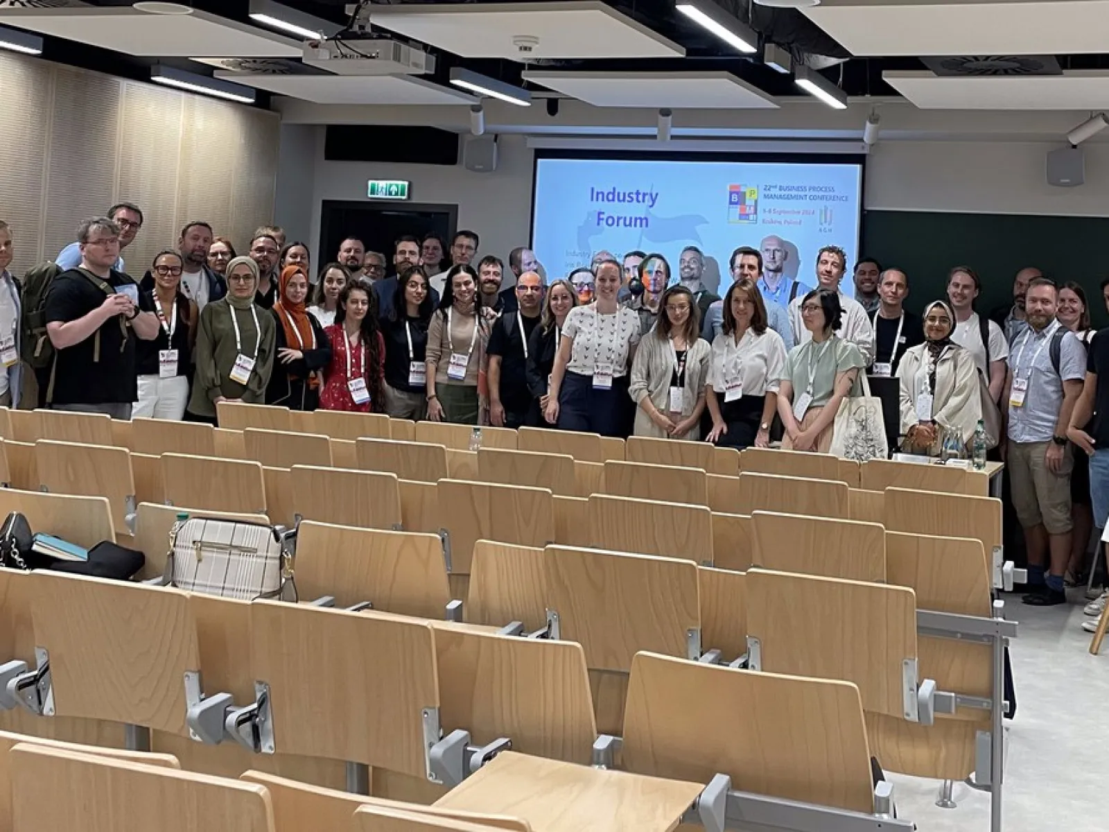 Eine Gruppe von Menschen steht in einem Hörsaal vor einer Präsentationsleinwand. Die Leinwand zeigt den Titel "Industry Forum". Die Personen lächeln in die Kamera. Im Vordergrund sind leere Sitzreihen zu sehen.