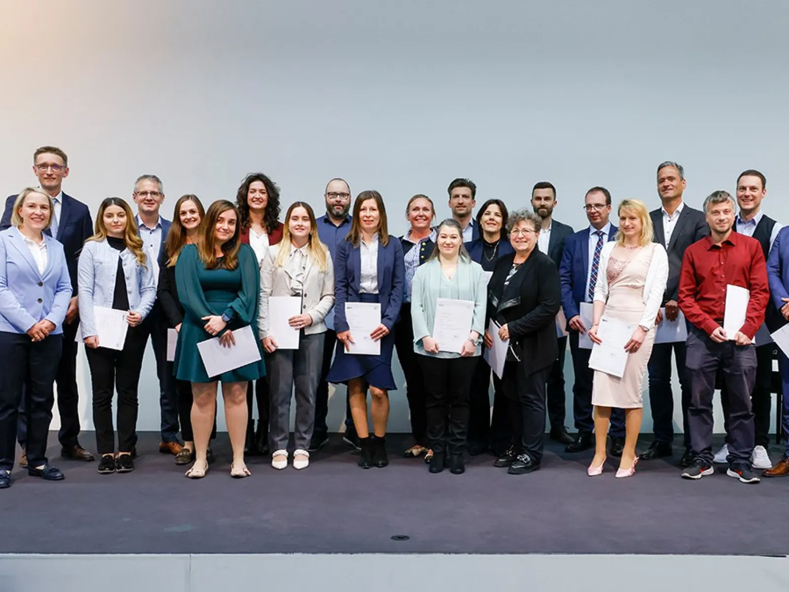Eine Gruppe von Absolventinnen und Absolventen steht auf einer Bühne und hält Zertifikate in den Händen. Sie lächeln in die Kamera. Die Personen sind formell gekleidet, und im Hintergrund ist eine neutrale Wand zu sehen.