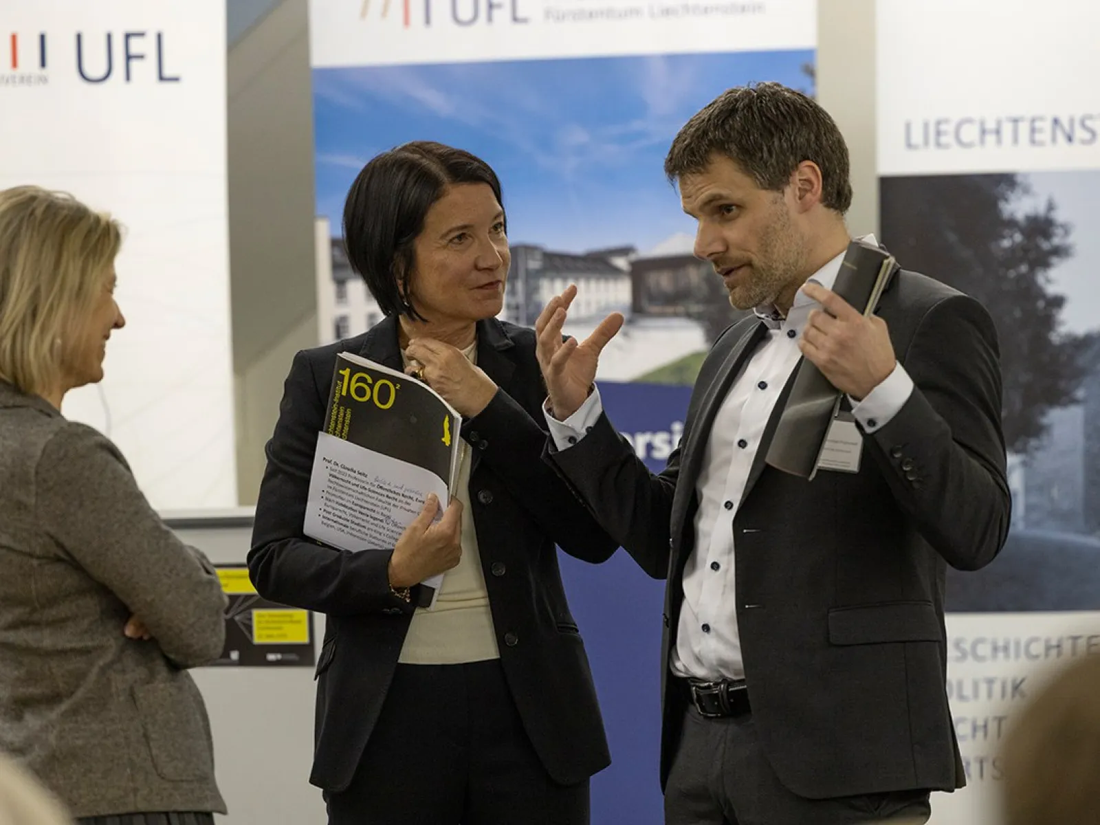 Drei Personen stehen in einem Gespräch vor Roll-Up-Bannern der Universität Liechtenstein und des Liechtenstein-Instituts. Eine Person hält ein Buch in der Hand. Die Szene findet in einem formellen Rahmen statt, vermutlich bei einer akademischen Veranstaltung.
