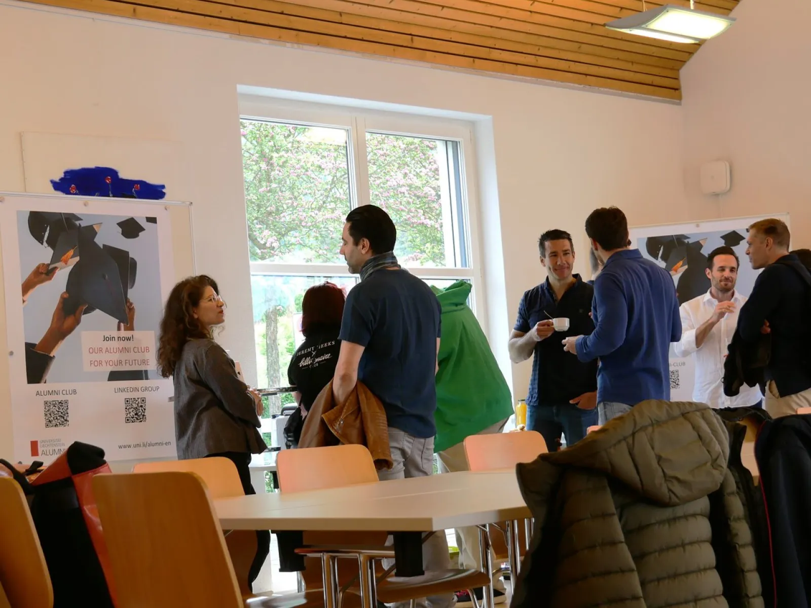 Eine Gruppe von Menschen steht in einem Raum der Universität Liechtenstein und unterhält sich. Im Hintergrund sind Poster und ein Fenster zu sehen. Die Atmosphäre wirkt entspannt und gesellig.