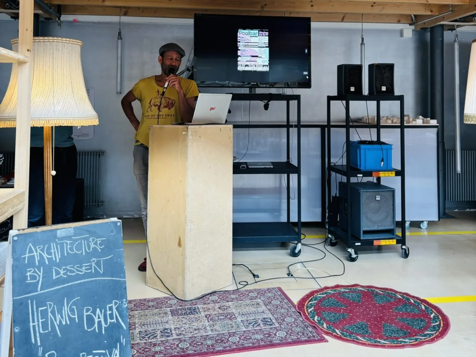 Ein Mann steht an einem Rednerpult in einem Raum mit Holzdach. Neben ihm ist ein Bildschirm mit einer Präsentation und ein Schild mit der Aufschrift „Architecture by Dessert, Herwig Bauer, Poolbarfestival“. Der Raum ist mit Teppichen und Stehlampen dekoriert.