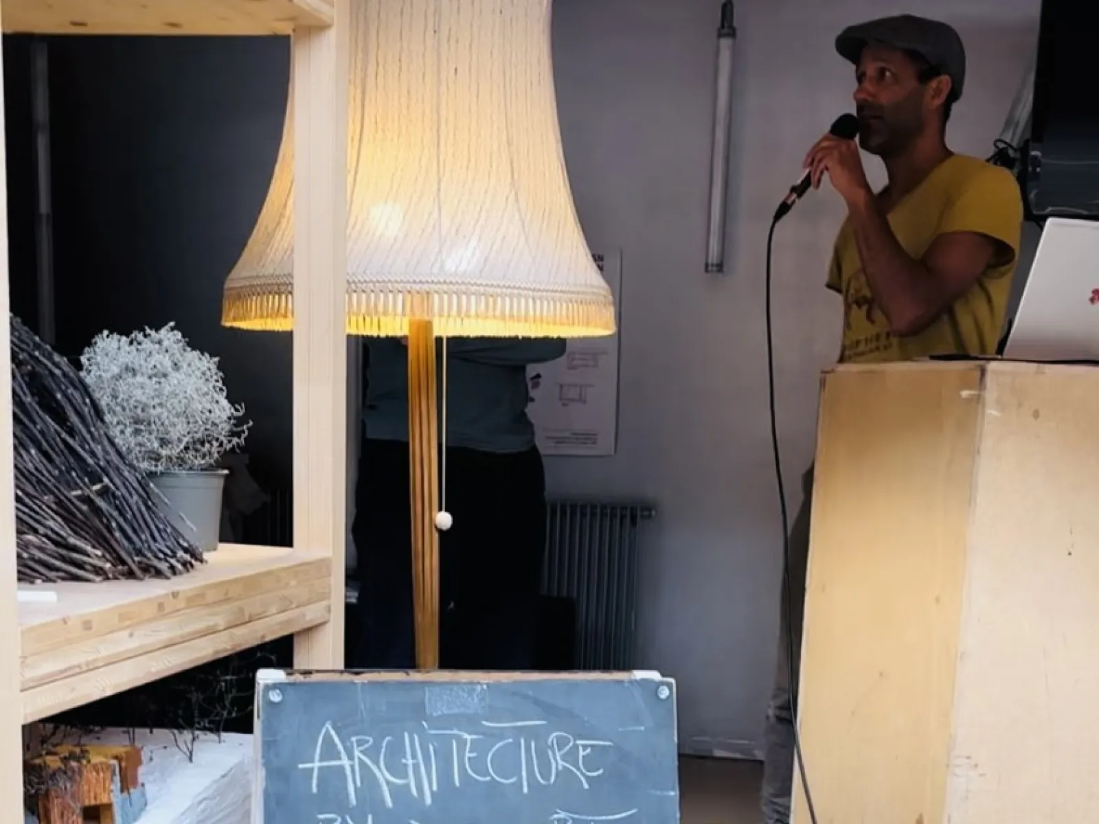 Ein Mann spricht in ein Mikrofon neben einem Rednerpult. Im Vordergrund steht eine Tafel mit der Aufschrift „Architecture by Dessert, Herwig Bauer, Poolbarfestival“. Links sind ein Regal und eine Stehlampe zu sehen.