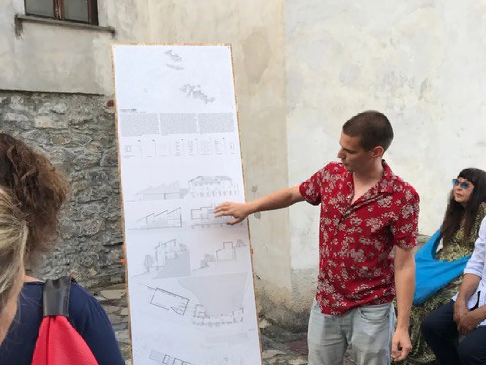 Ein Student präsentiert architektonische Skizzen auf einem großen Plakat in einem historischen Dorf. Um ihn herum stehen und sitzen mehrere Personen, die aufmerksam zuhören. Die Szene spielt sich in einem gepflasterten Außenbereich ab.