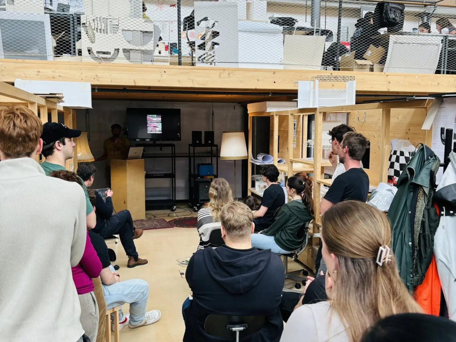 Eine Gruppe von Studierenden sitzt in einem Architekturatelier und hört einem Vortrag zu. Im Hintergrund sind Regale und Arbeitsmaterialien zu sehen.