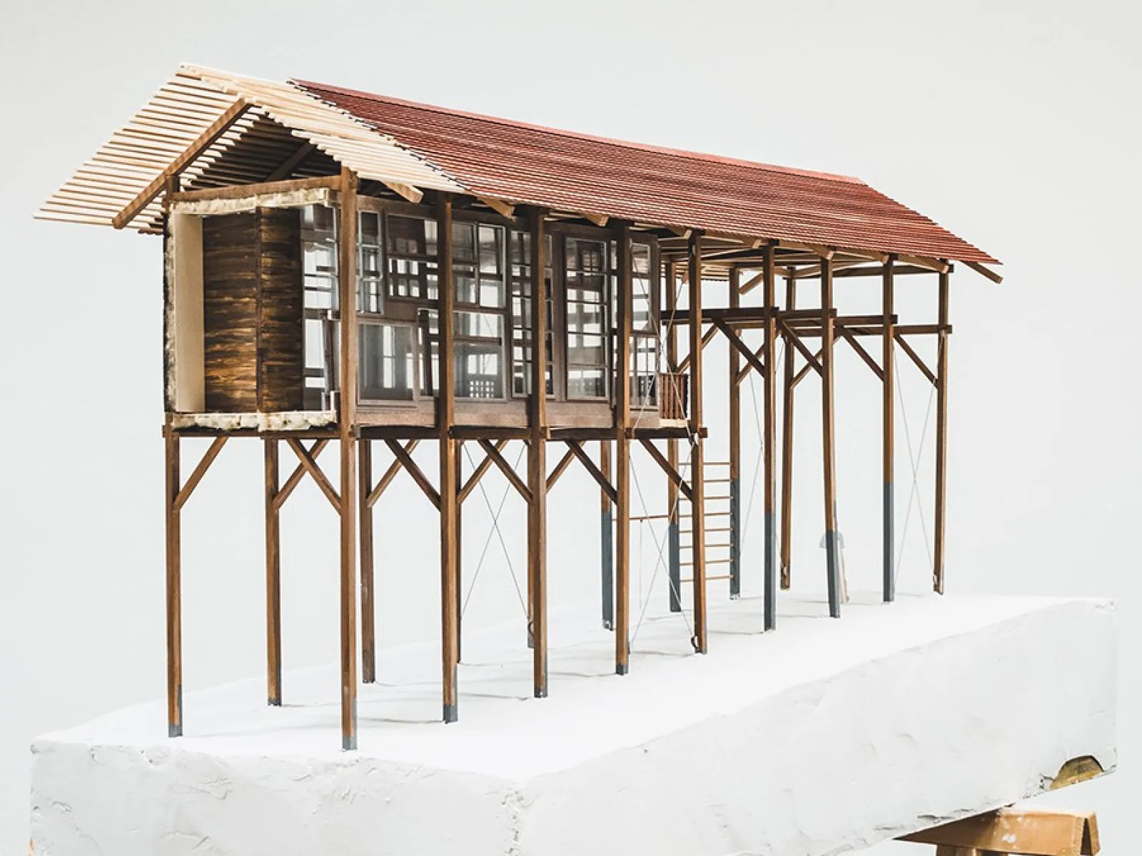 Ein Architekturmodell eines Strickbaus auf Stelzen, das auf einem weißen Sockel präsentiert wird. Das Modell zeigt eine lineare Struktur mit einem Satteldach und großen Fenstern, die traditionelle Bauweise und moderne Elemente kombinieren.