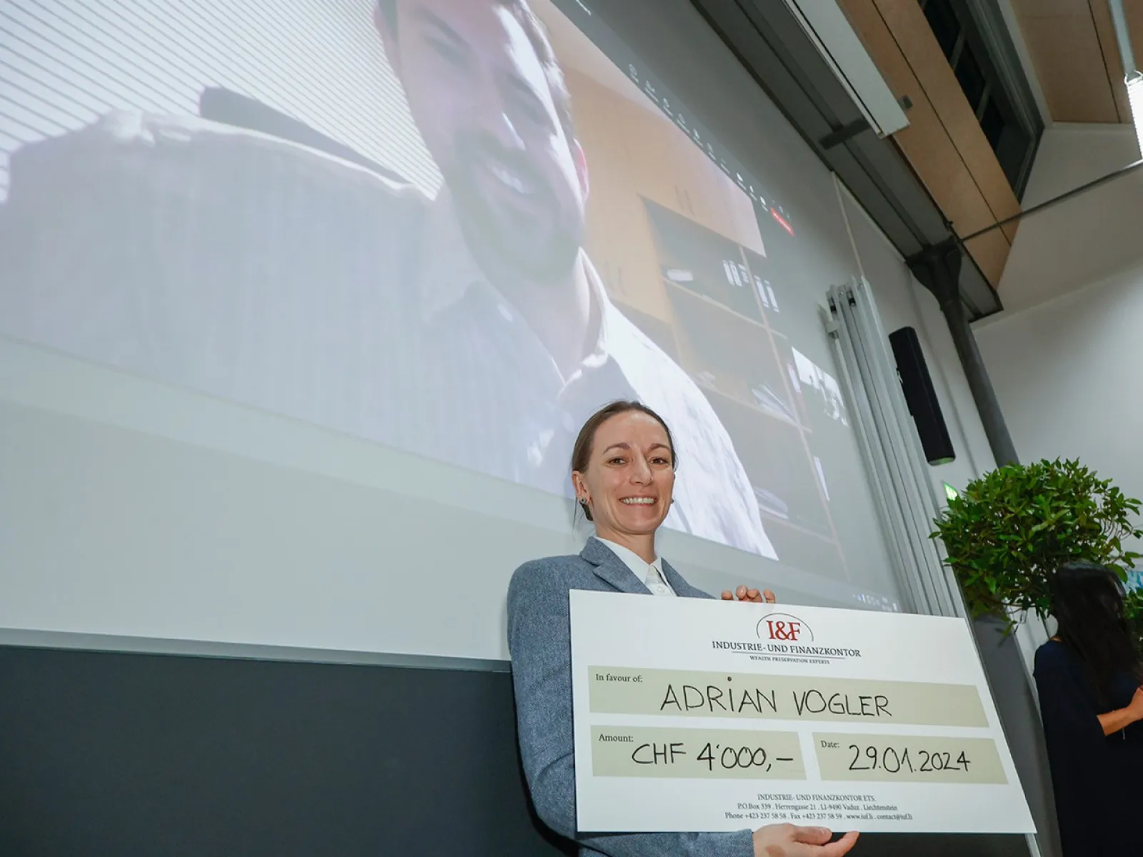 Eine Person hält einen großen Scheck mit der Aufschrift "Adrian Vogler, CHF 4,000, 29.01.2024" in einem Vortragsraum. Im Hintergrund ist eine Projektion eines lächelnden Mannes auf einer Leinwand zu sehen.