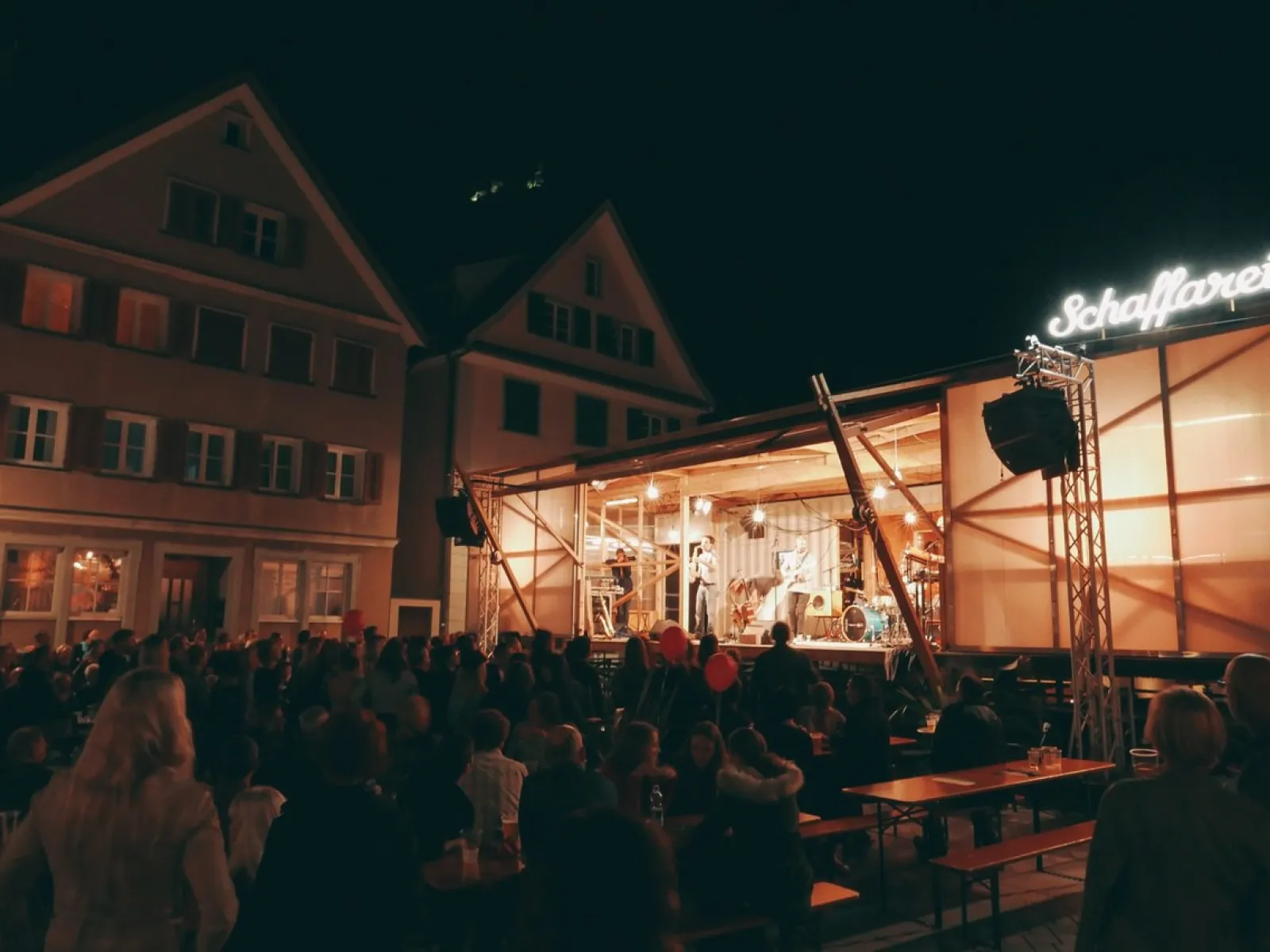 Eine beleuchtete Bühne des Base Camps der Universität Liechtenstein bei Nacht, umgeben von einem Publikum. Die Bühne ist modular und mobil, passend zur temporären Nutzung für Veranstaltungen und Stadtentwicklungsprojekte.