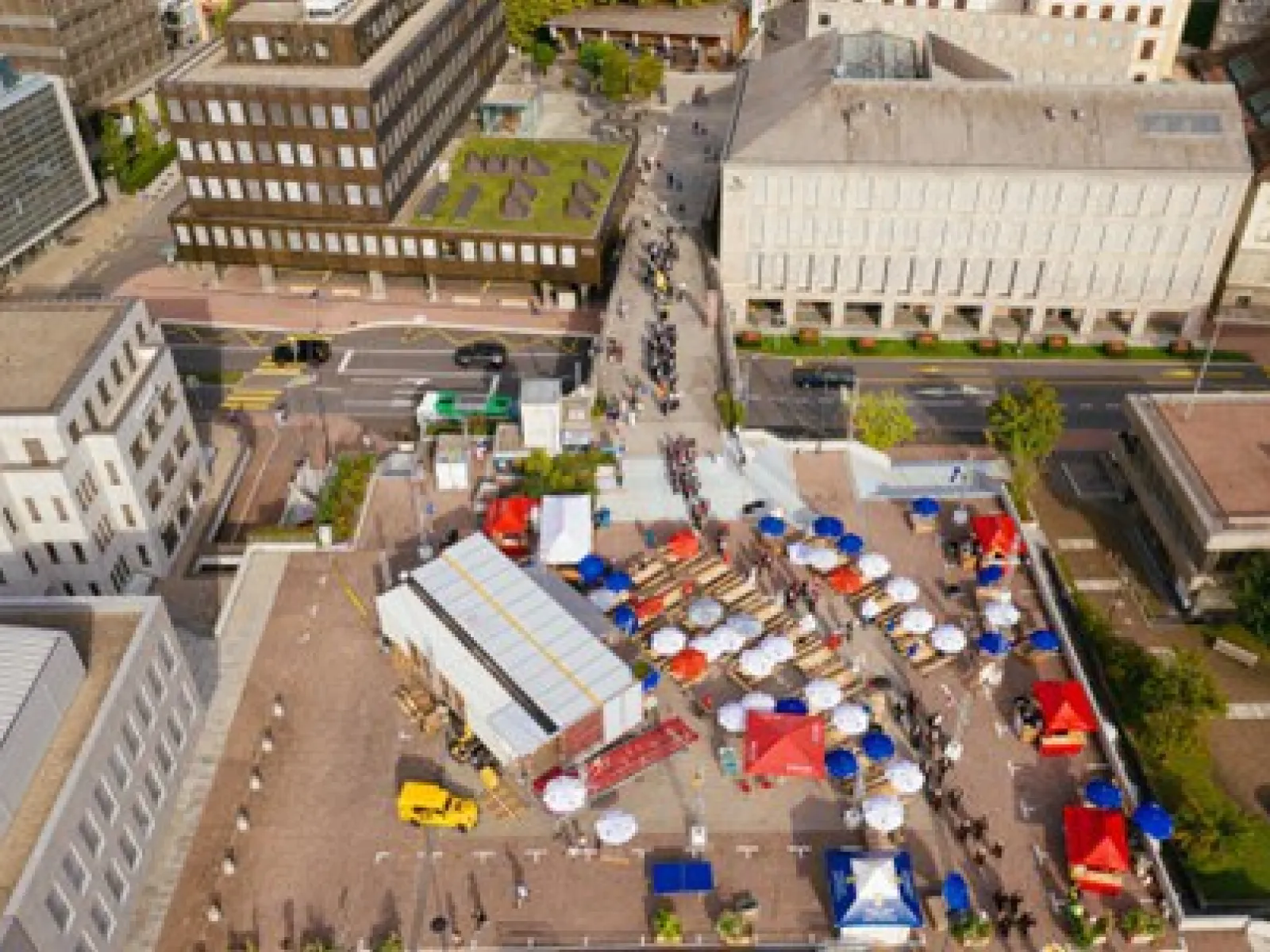 Luftaufnahme eines städtischen Platzes mit dem Base Camp der Universität Liechtenstein. Zu sehen sind modulare Strukturen, bunte Zelte und Menschen, die sich auf dem Platz bewegen. Gebäude umgeben den Platz, und eine Straße verläuft im Hintergrund.