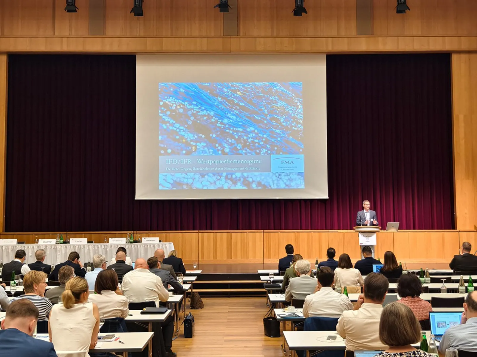 Ein Vortragssaal mit Teilnehmern, die an Tischen sitzen und einem Redner auf der Bühne zuhören. Eine große Leinwand zeigt eine Präsentation. Der Raum ist gut besucht und die Atmosphäre wirkt professionell.
