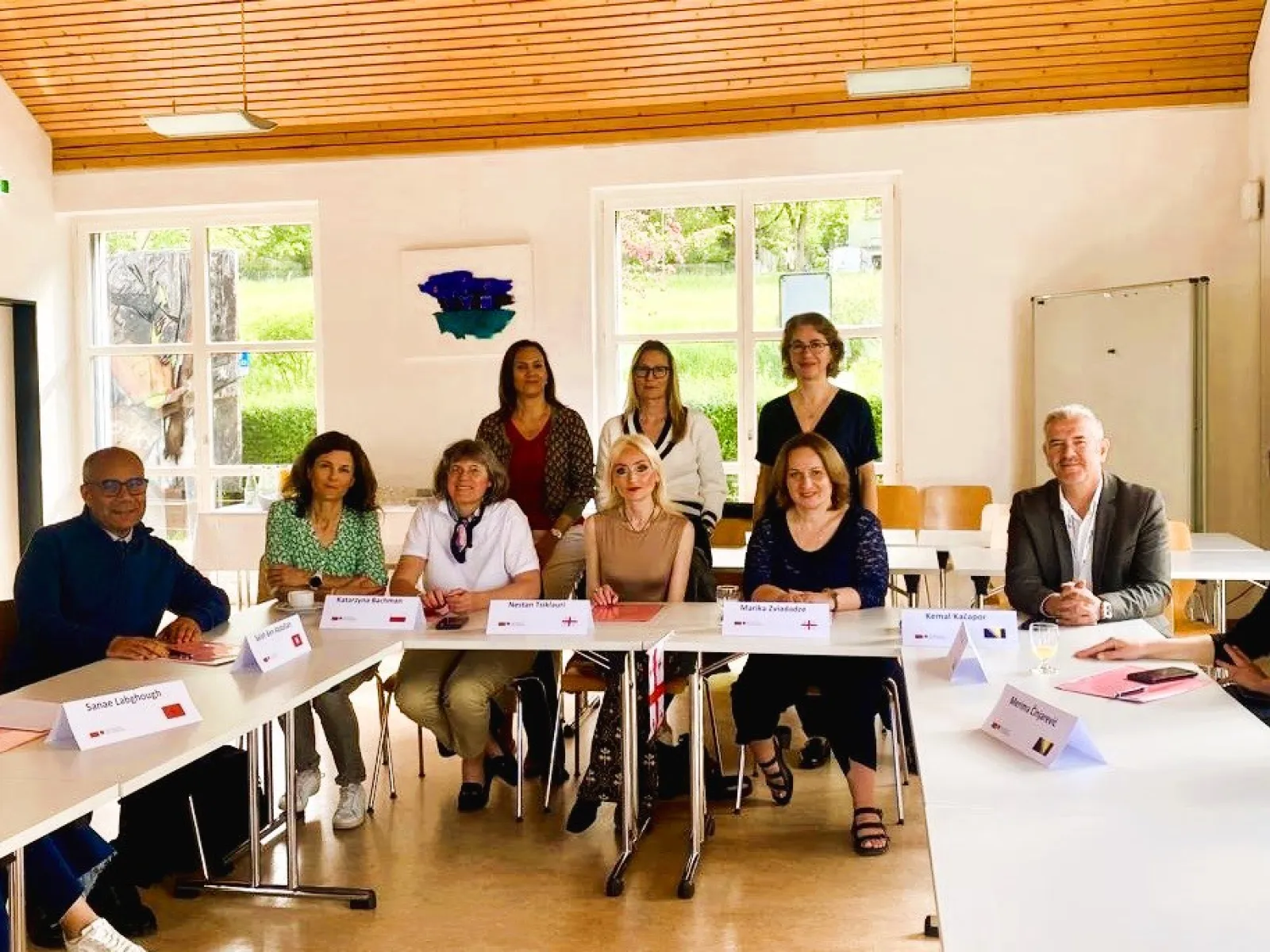 Eine Gruppe von zwölf Personen sitzt an einem U-förmigen Tisch in einem hellen Raum. Sie sind Teil eines internationalen Besuchsprogramms an der Universität Liechtenstein. Im Hintergrund sind große Fenster und eine Holzbalkendecke zu sehen.