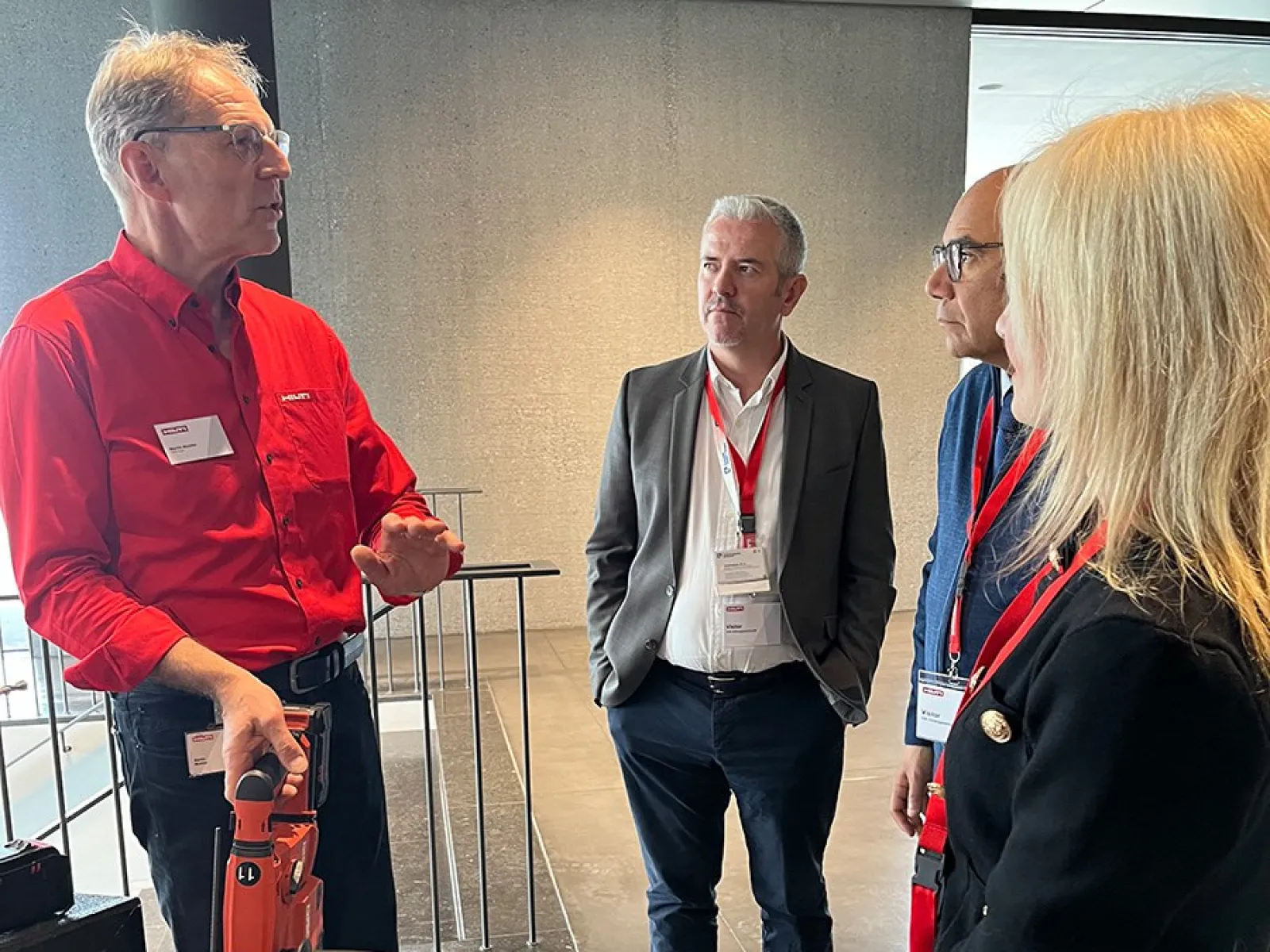 Eine Gruppe von vier Personen steht in einem modernen Gebäude. Eine Person hält ein Werkzeug und erklärt etwas den anderen. Alle tragen Namensschilder und hören aufmerksam zu.