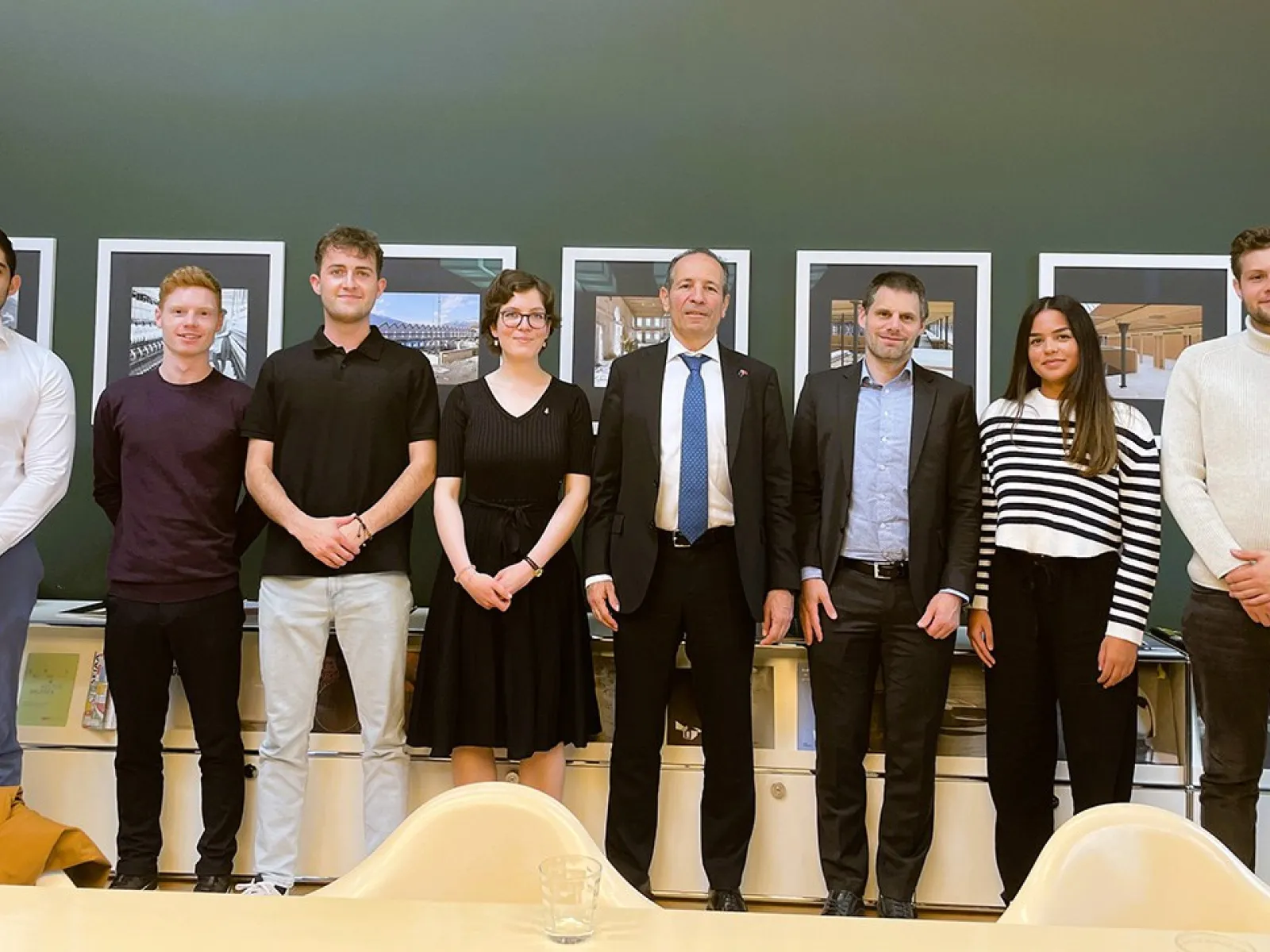 Eine Gruppe von sieben Personen, darunter Studierende und zwei Männer in Anzügen, steht lächelnd vor einer Wand mit Bildern. Sie befinden sich in einem Raum der Universität Liechtenstein.