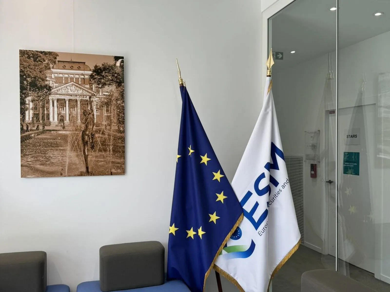 Ein moderner Warteraum mit blauen Sitzmöbeln, zwei Flaggen – eine der Europäischen Union und eine mit dem Logo des Europarats – und einem gerahmten Bild eines historischen Gebäudes an der Wand.