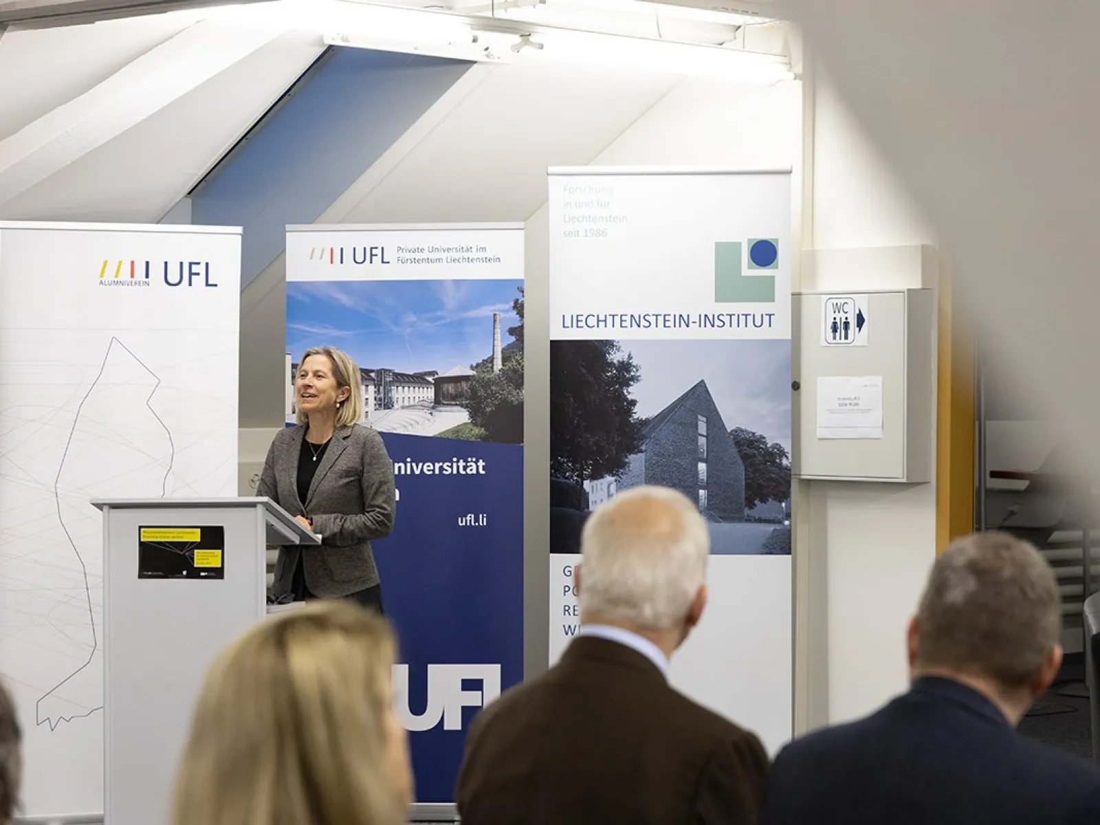 Eine Frau steht an einem Rednerpult vor Bannern der Universität Liechtenstein, der UFL und des Liechtenstein-Instituts. Im Vordergrund sind unscharf einige Zuhörer zu sehen.