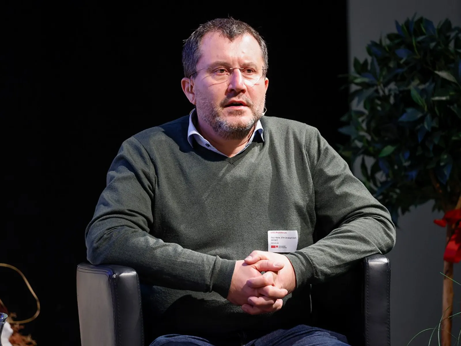 Eine Person sitzt auf einem schwarzen Sessel und hält die Hände gefaltet. Im Hintergrund sind Pflanzen zu sehen.