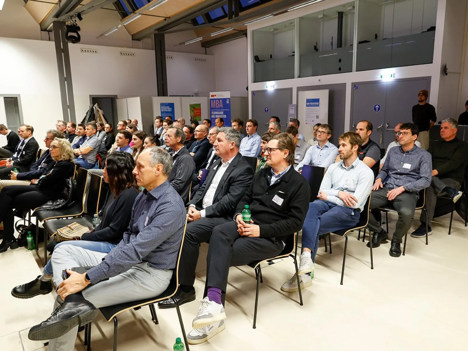 Eine Gruppe von Menschen sitzt in einem modernen Konferenzraum der Universität Liechtenstein. Sie hören aufmerksam einem Vortrag zu, der sich mit den Herausforderungen und Chancen der Nachhaltigkeitsregulierung für kleine und mittelständische Unternehmen befasst.