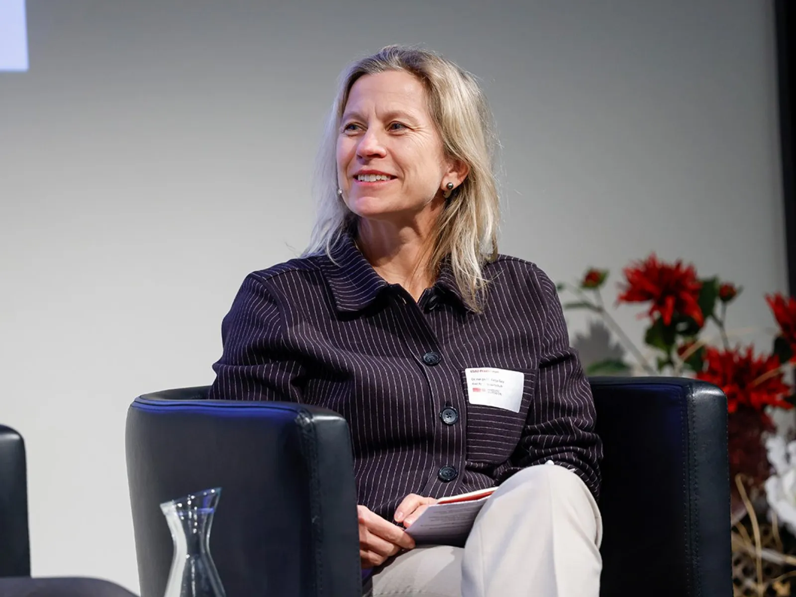 Eine Person sitzt lächelnd auf einem Podium in einem Konferenzraum. Im Hintergrund sind Blumenarrangements zu sehen.