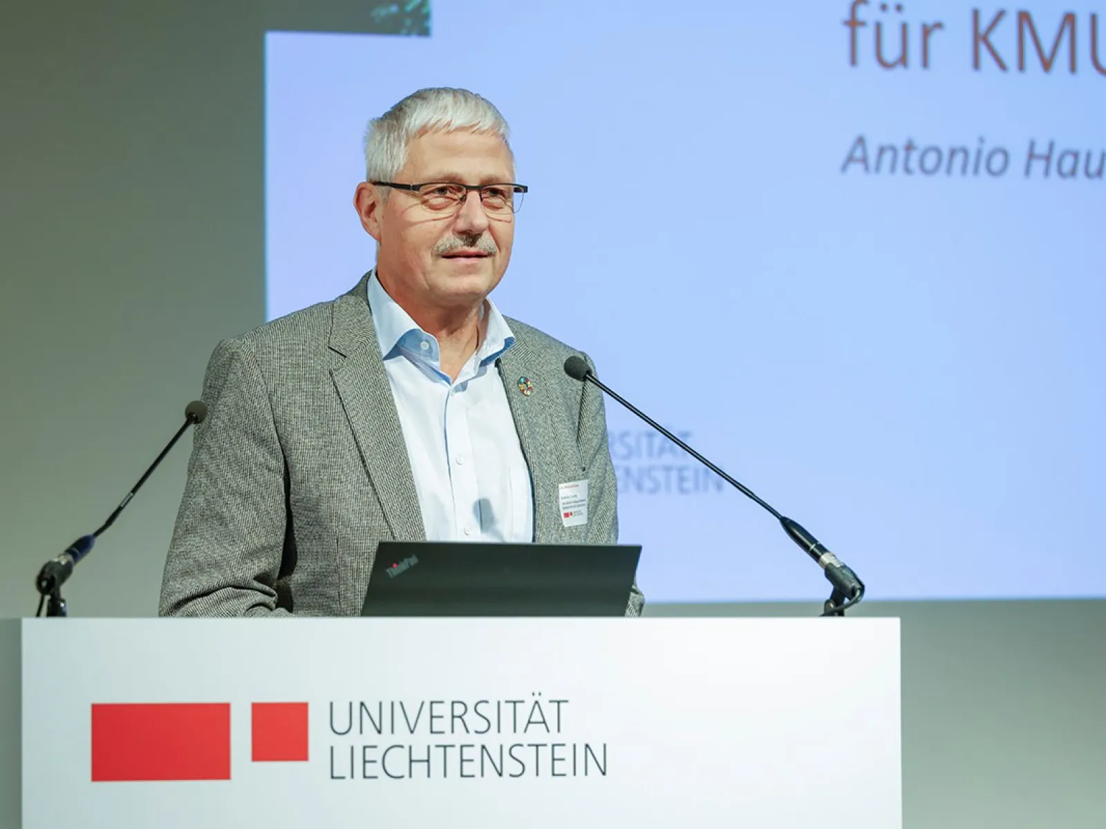 Ein Redner steht an einem Podium mit dem Logo der Universität Liechtenstein und spricht in ein Mikrofon. Im Hintergrund ist eine Präsentationsfolie zu sehen.
