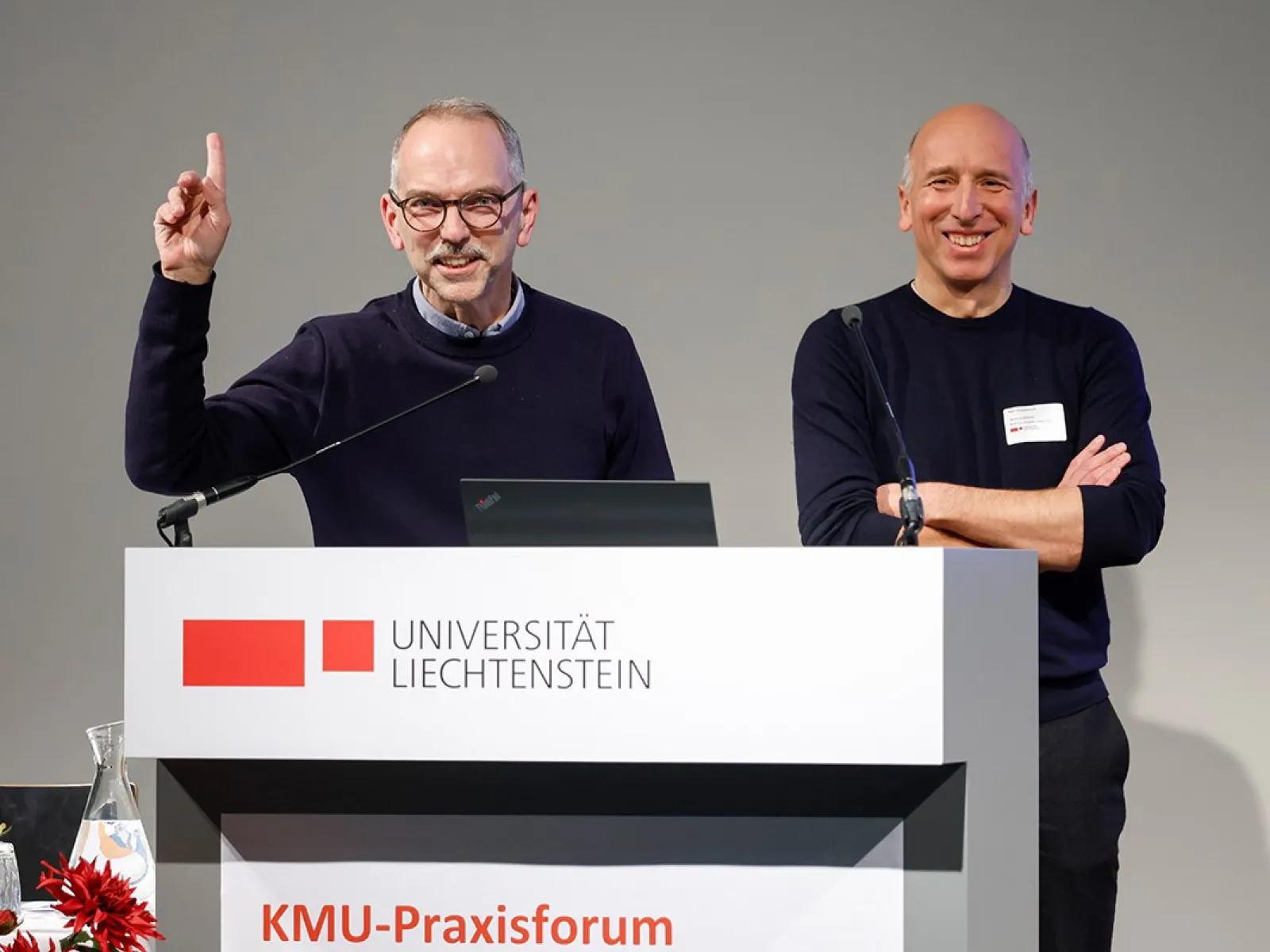 Zwei Männer stehen lächelnd hinter einem Rednerpult mit dem Logo der Universität Liechtenstein. Einer hebt die Hand, während der andere die Arme verschränkt. Im Vordergrund sind rote Blumen zu sehen.