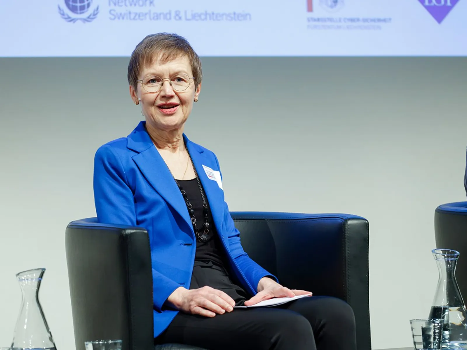 Eine Person in einem blauen Blazer sitzt auf einem Podium und spricht bei einer Veranstaltung an der Universität Liechtenstein. Im Hintergrund sind Logos von Partnerorganisationen zu sehen.