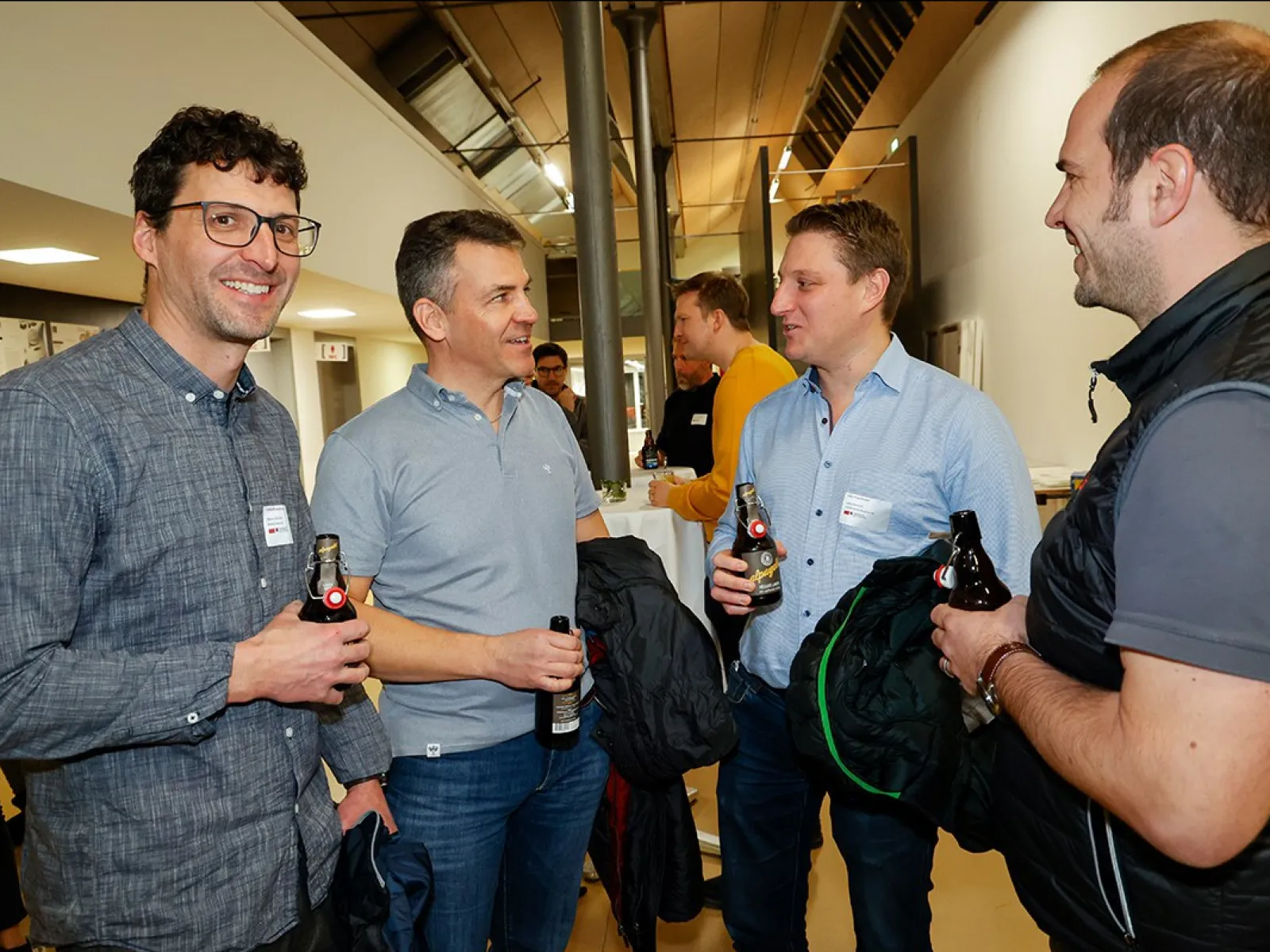 Vier Männer unterhalten sich bei einer Veranstaltung an der Universität Liechtenstein. Sie halten Getränke in der Hand und tragen Namensschilder. Die Atmosphäre wirkt entspannt und gesellig.