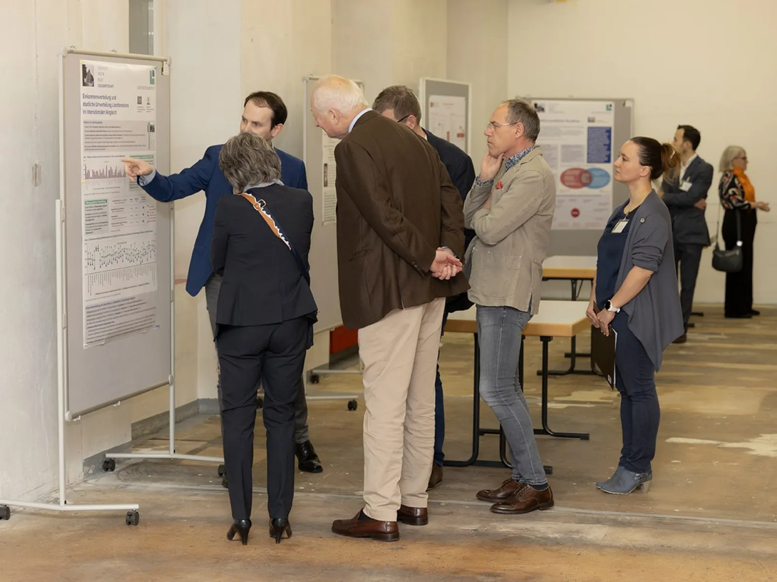 Eine Gruppe von Personen betrachtet eine wissenschaftliche Posterausstellung in einem Innenraum. Ein Mann zeigt auf ein Poster und erklärt den Inhalt. Im Hintergrund sind weitere Poster und Personen zu sehen.