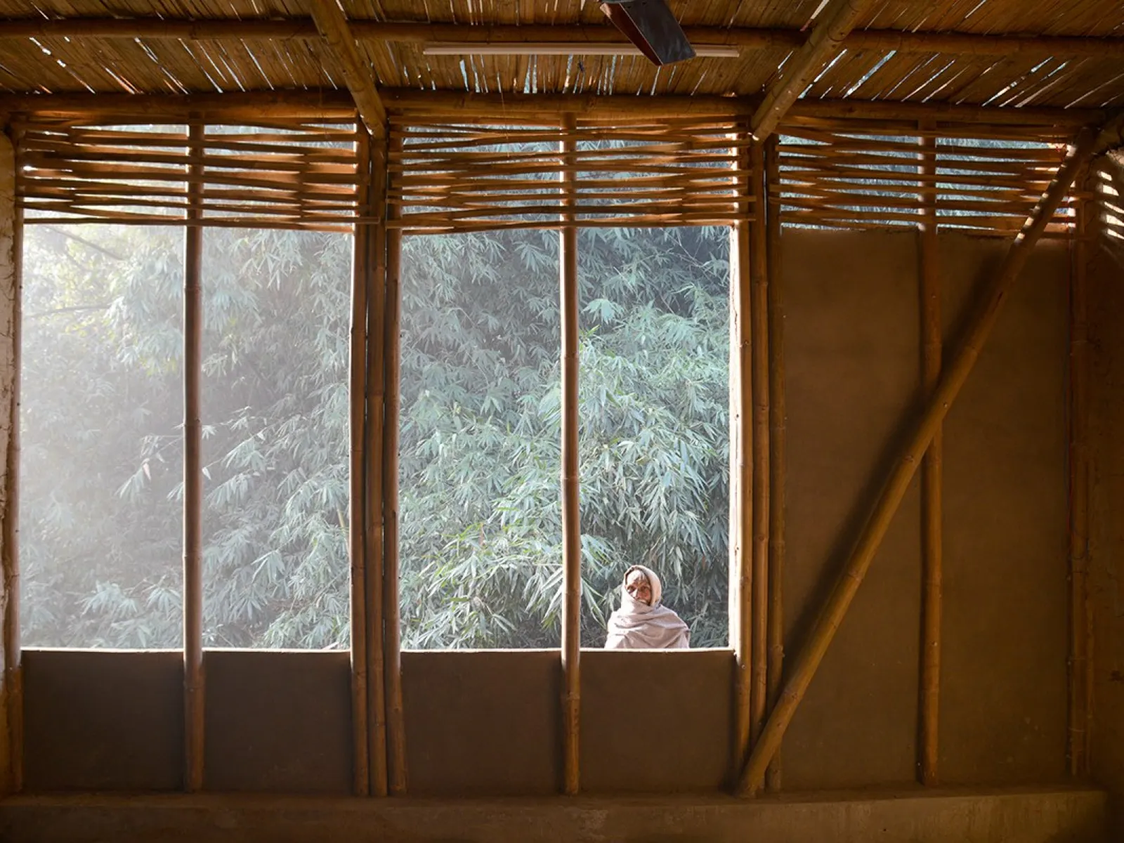 Innenansicht eines Gebäudes mit Wänden aus Lehm und Bambus, durch ein großes Fenster fällt Sonnenlicht auf eine Person, die draußen steht. Im Hintergrund sind Bäume zu sehen.