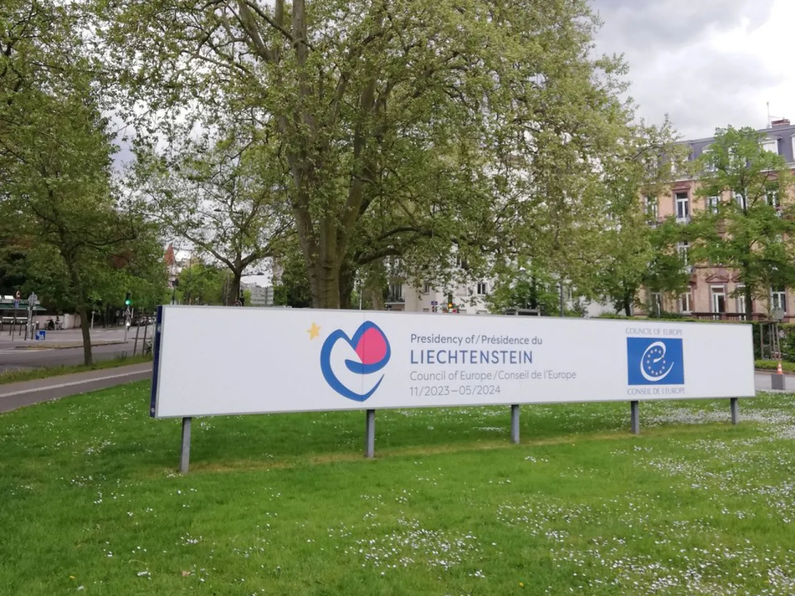 Ein großes Schild mit dem Logo des Fürstentums Liechtenstein und dem Europarat steht auf einer grünen Wiese, umgeben von Bäumen und Gebäuden im Hintergrund.