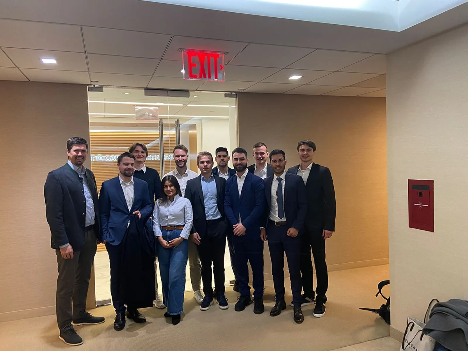 Eine Gruppe von Studierenden in Business-Kleidung steht in einem modernen Bürogebäude. Im Hintergrund ist eine Glastür zu sehen. Die Szene wirkt professionell und vermittelt den Eindruck einer Exkursion oder eines geschäftlichen Treffens.