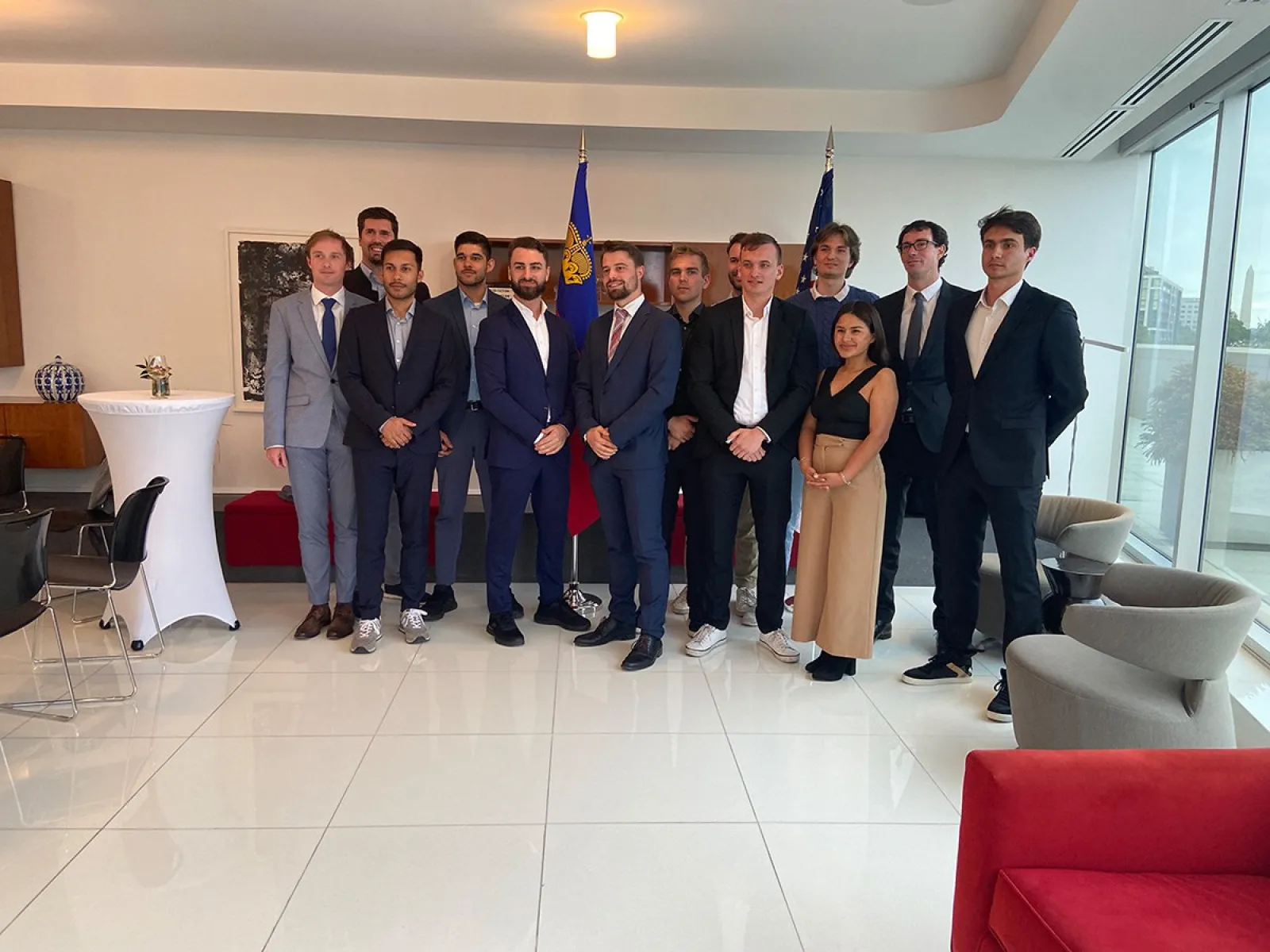 Gruppe von Studierenden und Begleitpersonen in Businesskleidung steht in einem modernen, hellen Raum mit Flaggen im Hintergrund.