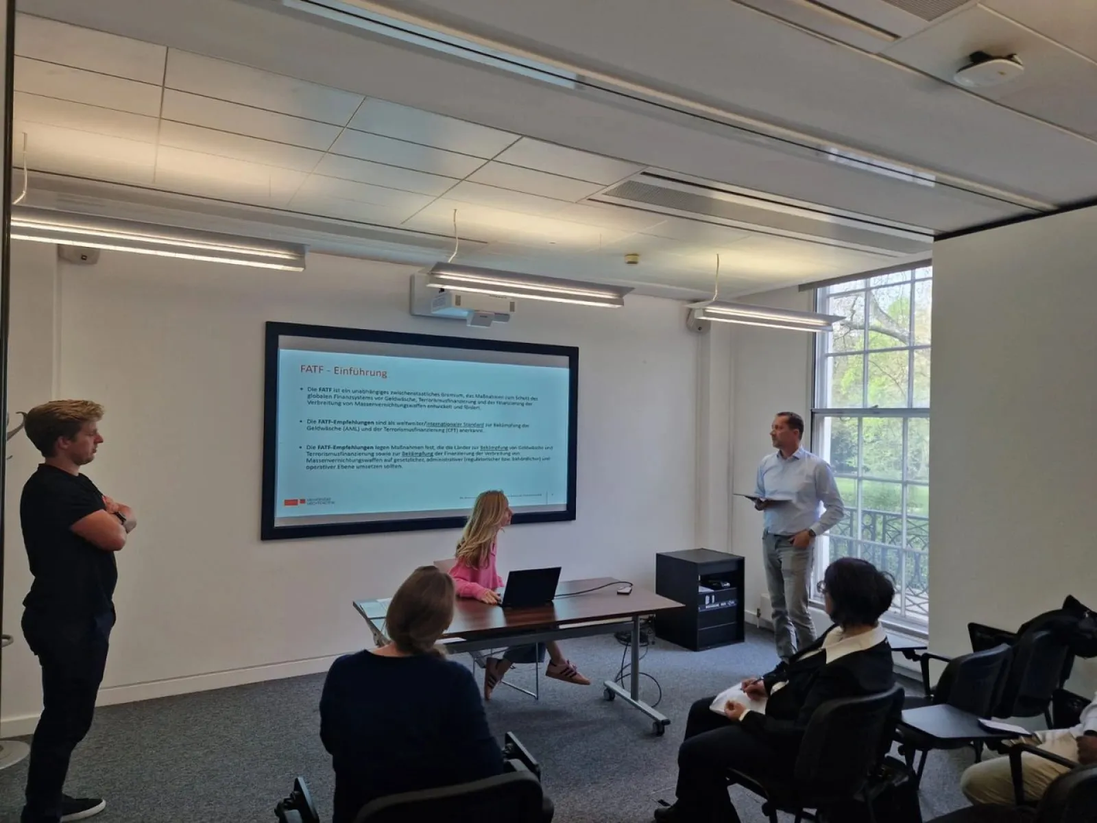 Studierende und Dozenten in einem Seminarraum der Universität Liechtenstein. Eine Präsentation wird auf einem großen Bildschirm gezeigt, während ein Dozent spricht und Studierende aufmerksam zuhören. Helle, moderne Umgebung mit großen Fenstern.