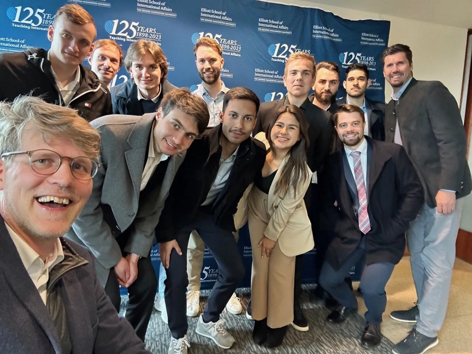 Gruppe von Studierenden und Dozenten der Universität Liechtenstein posiert vor einem Banner der Elliott School of International Affairs während einer Studienreise in den USA.