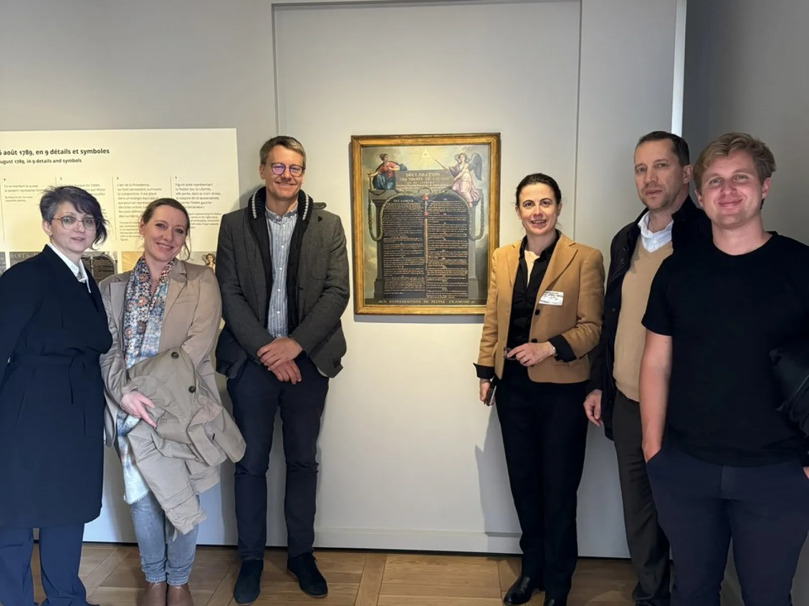 Eine Gruppe von sechs Personen steht in einem Ausstellungsraum vor einem Gemälde. Sie lächeln in die Kamera. Der Raum hat helle Wände und einen Holzboden.