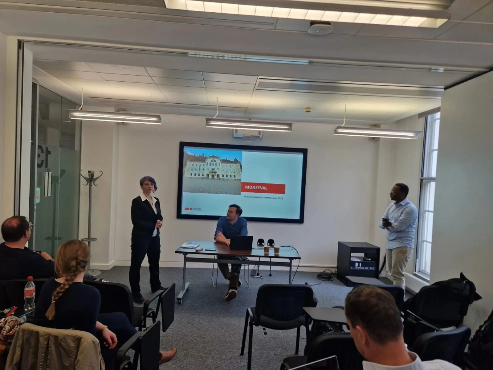 Ein Vortrag im Seminarraum: Eine Person steht links und spricht, während eine andere Person rechts steht. In der Mitte sitzt jemand an einem Tisch. Im Hintergrund ist eine Präsentation auf einem Bildschirm zu sehen. Zuschauer sitzen im Vordergrund.