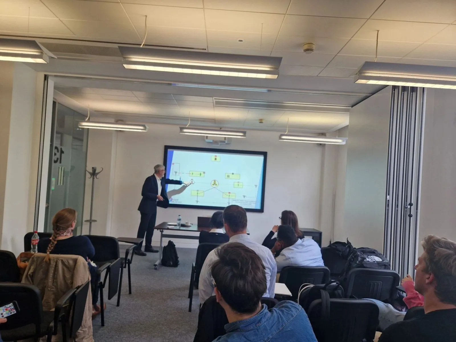 Ein Vortragender präsentiert vor einer Gruppe von Studierenden in einem modernen Seminarraum. Auf einem großen Bildschirm sind Diagramme zu sehen. Die Studierenden hören aufmerksam zu.