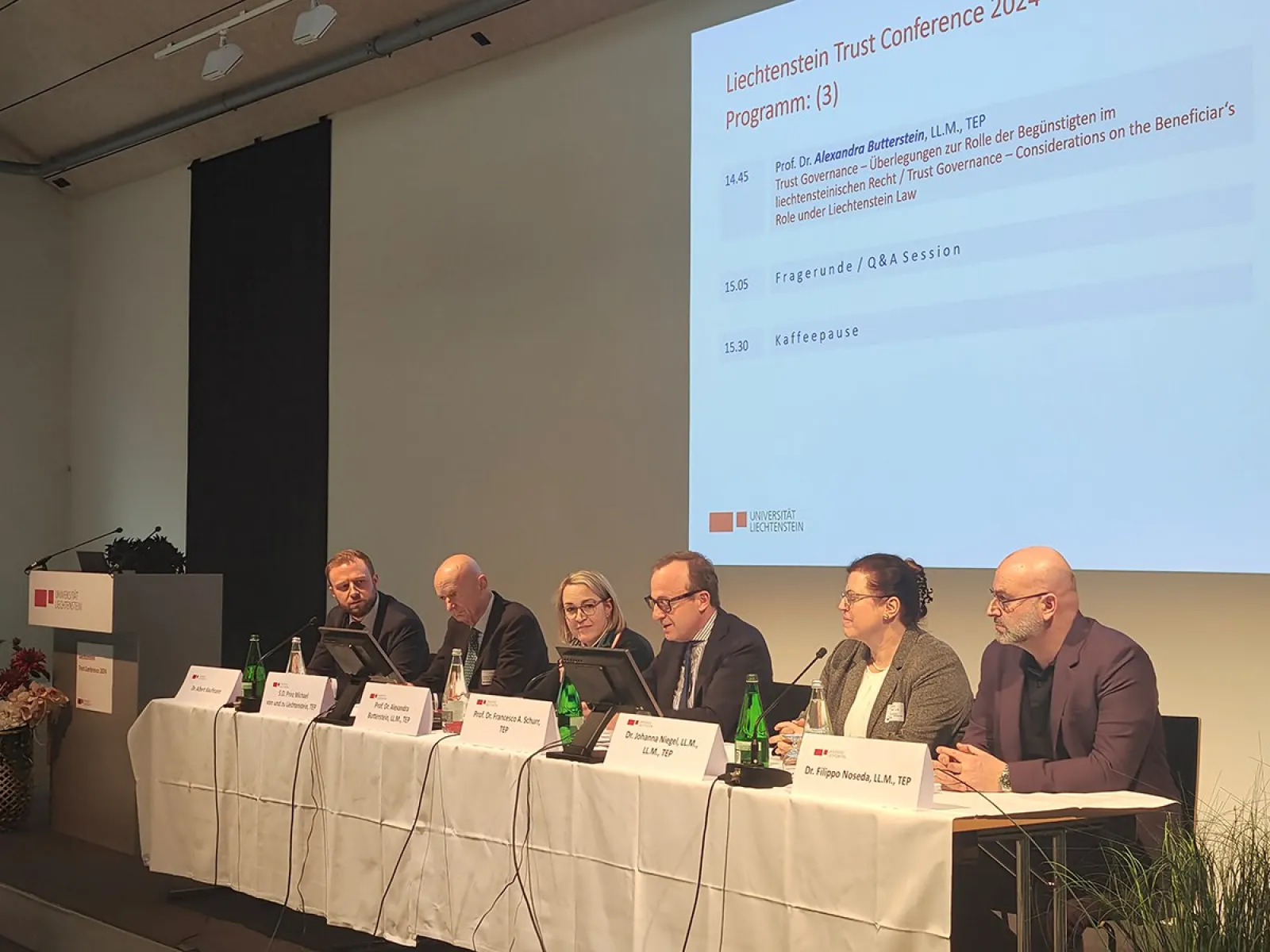 Ein Podium mit sechs Personen bei der Liechtenstein Trust Conference 2024. Sie sitzen an einem Tisch mit Mikrofonen und Namensschildern. Im Hintergrund ist eine Leinwand mit dem Konferenzprogramm zu sehen.