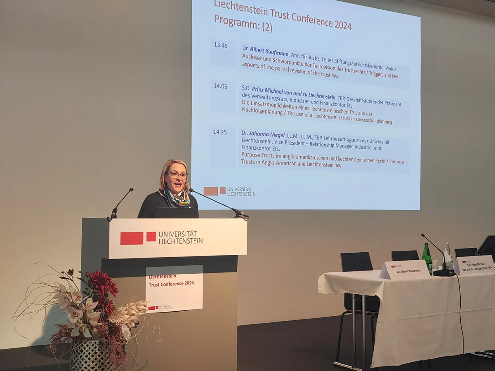 Eine Rednerin steht am Podium der Universität Liechtenstein während der Liechtenstein Trust Conference 2024. Im Hintergrund ist eine Präsentationsfolie mit dem Konferenzprogramm zu sehen. Ein Tisch mit Mikrofonen und Wasserflaschen befindet sich rechts im Bild.