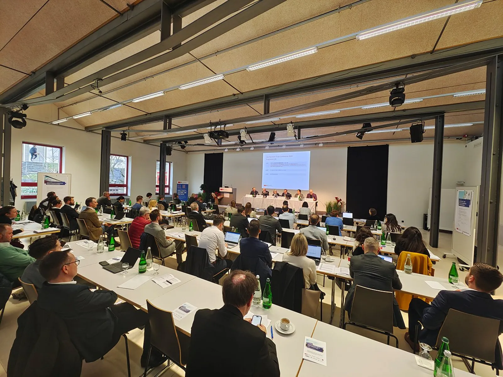 Ein Auditorium der Universität Liechtenstein, gefüllt mit Teilnehmenden der Liechtenstein Trust Conference 2024. Vorne eine Präsentation auf einer Leinwand, umgeben von Vortragenden. Die Teilnehmenden sitzen an Tischen mit Getränken und Notizen.