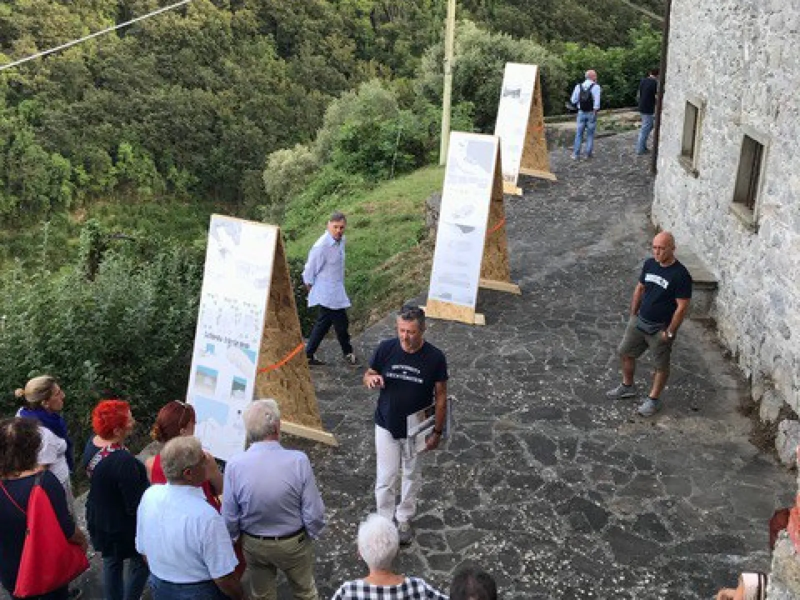 Eine Gruppe von Menschen betrachtet im Freien aufgestellte Tafeln mit Informationen. Die Szene spielt sich auf einem gepflasterten Weg neben einem alten Steingebäude ab, umgeben von üppiger, grüner Landschaft.