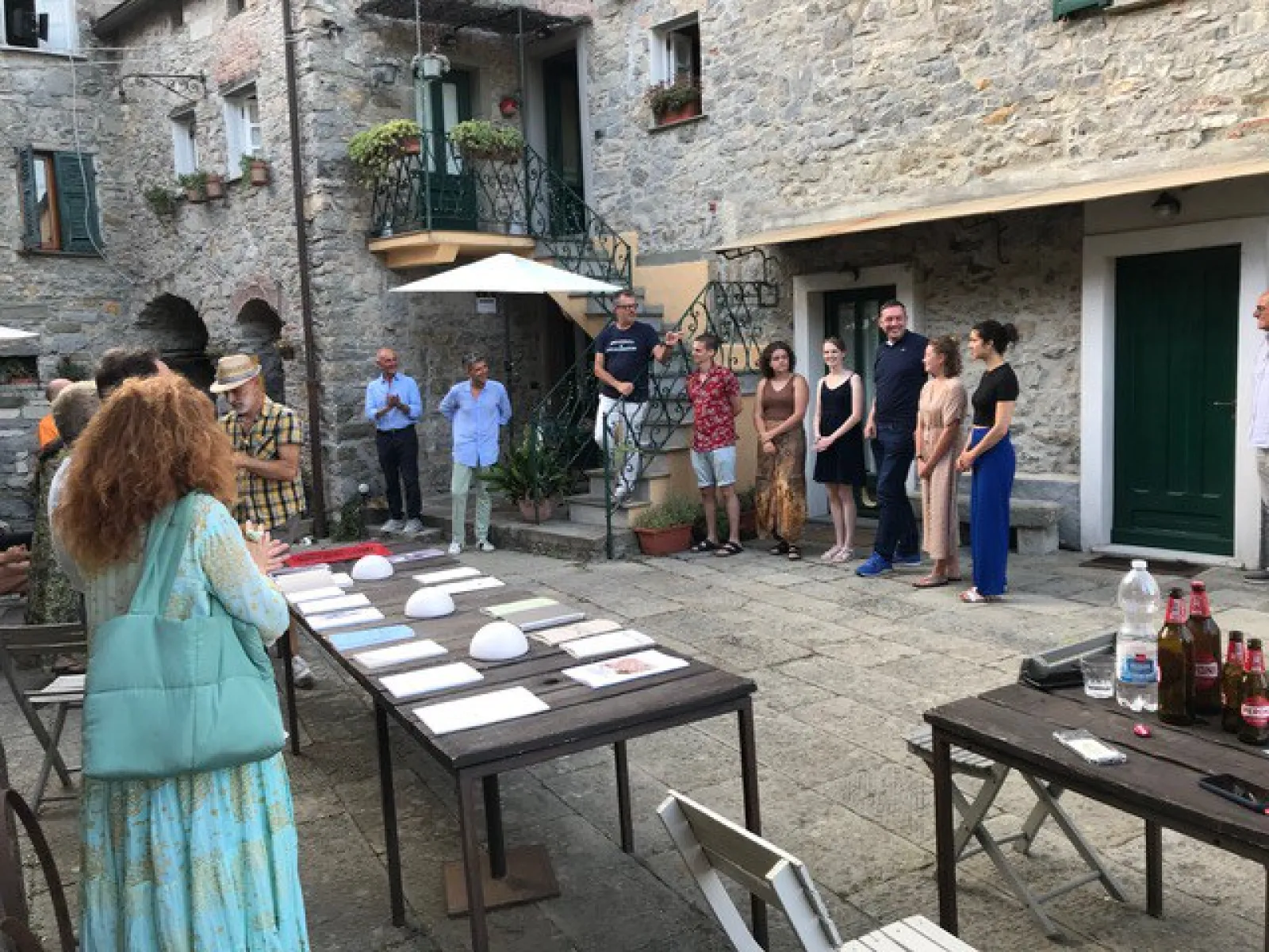 Eine Gruppe von Menschen versammelt sich in einem historischen Innenhof eines Dorfes. Auf Tischen liegen Dokumente und Objekte aus. Die Szene zeigt eine Präsentation oder Diskussion im Freien, umgeben von alten Steinmauern und Pflanzen.