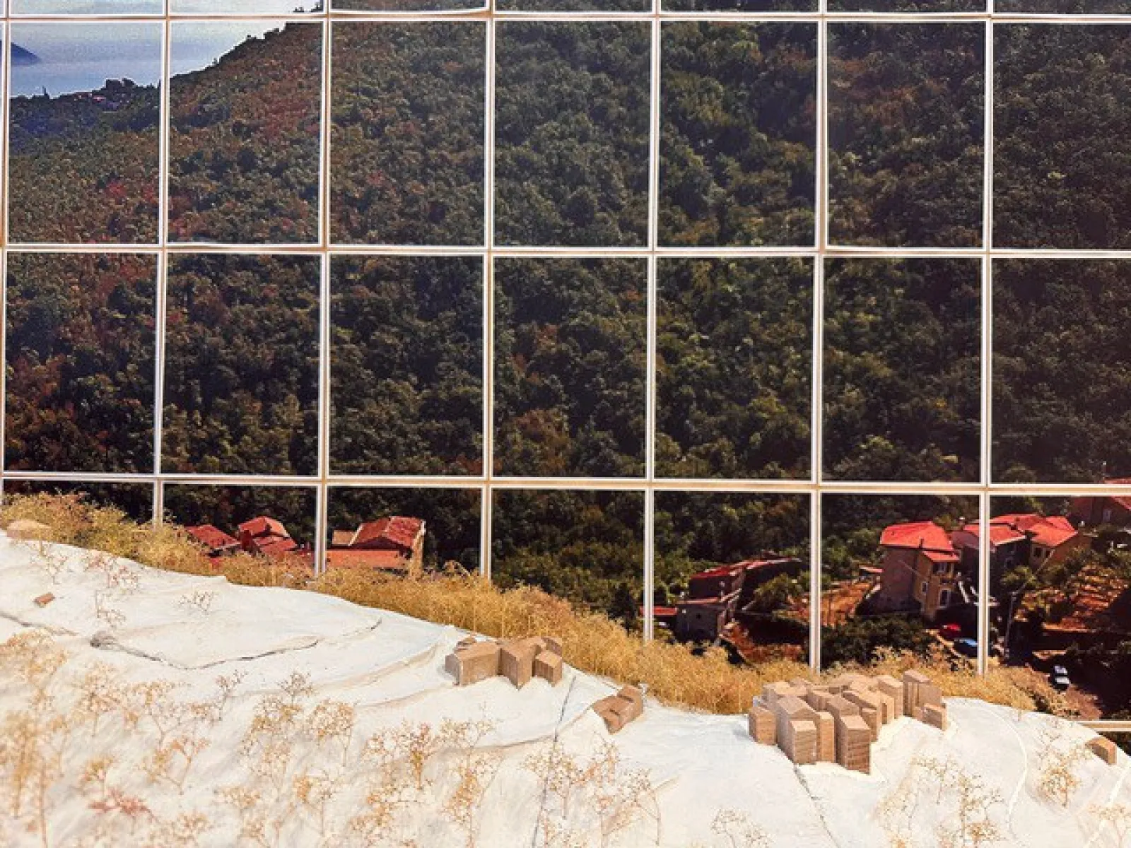 Ein Modell zeigt eine hügelige Landschaft mit kleinen Gebäuden und Vegetation, im Hintergrund ist ein großes Foto einer bewaldeten Berglandschaft zu sehen.