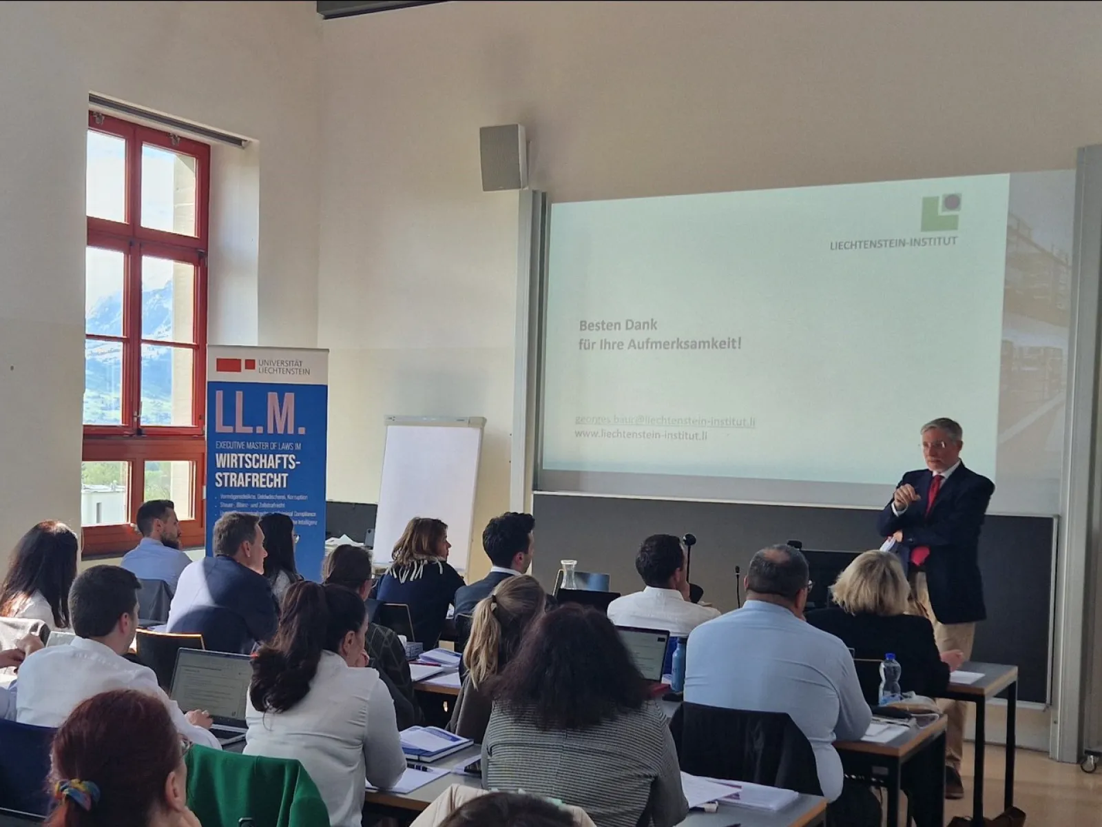 Ein Vortragender steht vor einer Gruppe von Teilnehmenden in einem Seminarraum der Universität Liechtenstein. Auf der Leinwand ist eine Präsentationsfolie mit dem Text "Besten Dank für Ihre Aufmerksamkeit!" zu sehen. Im Hintergrund ist ein Banner mit der Aufschrift "LL.M. Wirtschafts-Strafrecht" sichtbar.