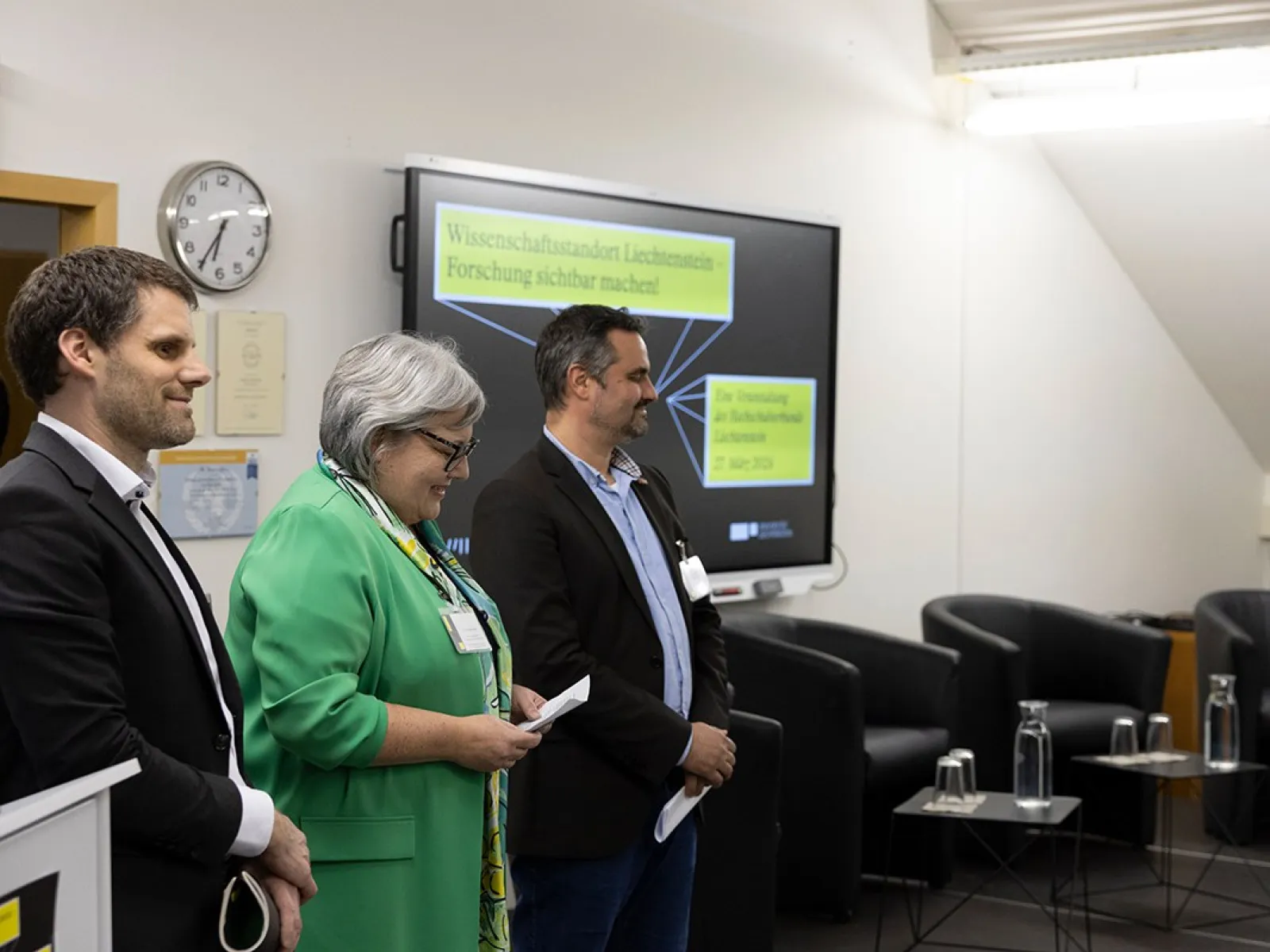 Drei Personen stehen nebeneinander in einem Konferenzraum. Im Hintergrund ist ein Bildschirm mit der Aufschrift "Wissenschaftsverbund Liechtenstein" zu sehen. Links im Bild ist ein Rednerpult, rechts sind leere Stühle und Tische. Die Personen tragen Namensschilder und lächeln.