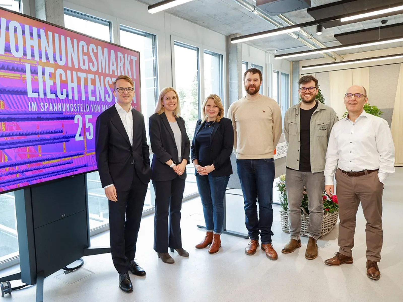 Gruppe von sechs Personen steht vor einem Bildschirm mit der Aufschrift "Wohnungsmarkt Liechtenstein". Die Personen lächeln und sind in einem modernen Raum mit großen Fenstern.