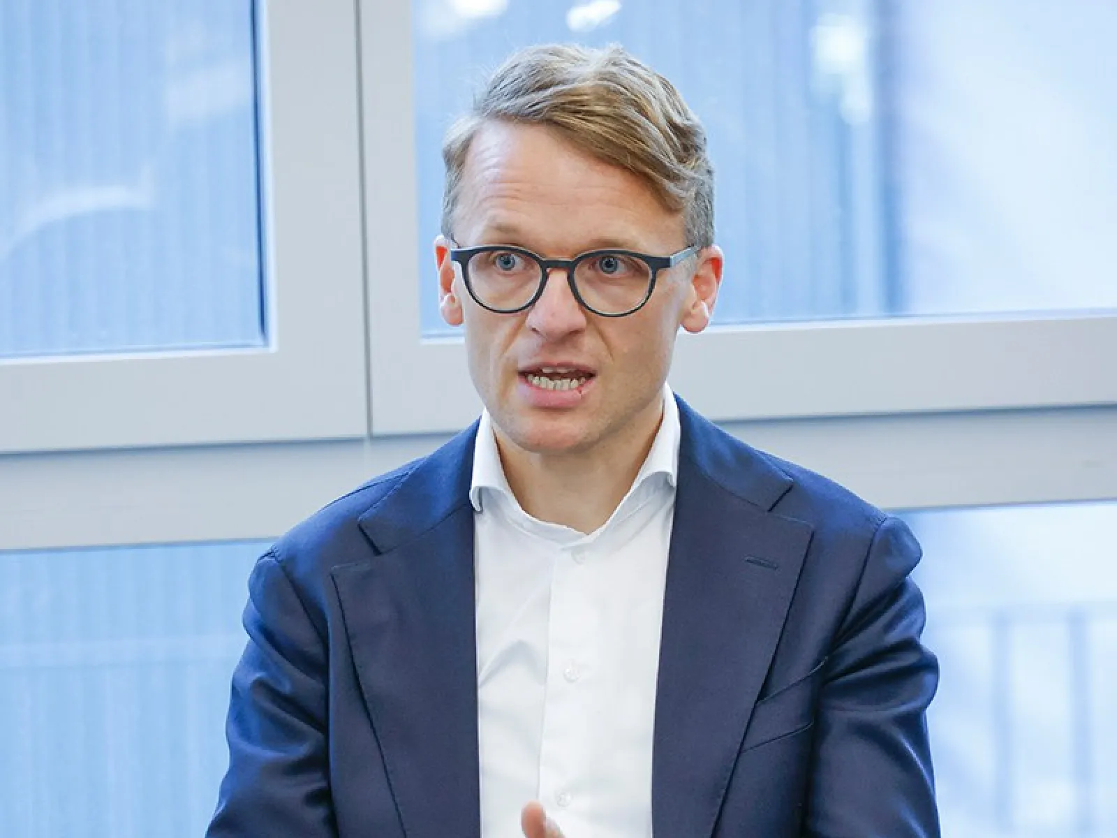 Ein Mann in einem Anzug spricht gestikulierend während einer Podiumsdiskussion in einem modernen Raum mit großen Fenstern.